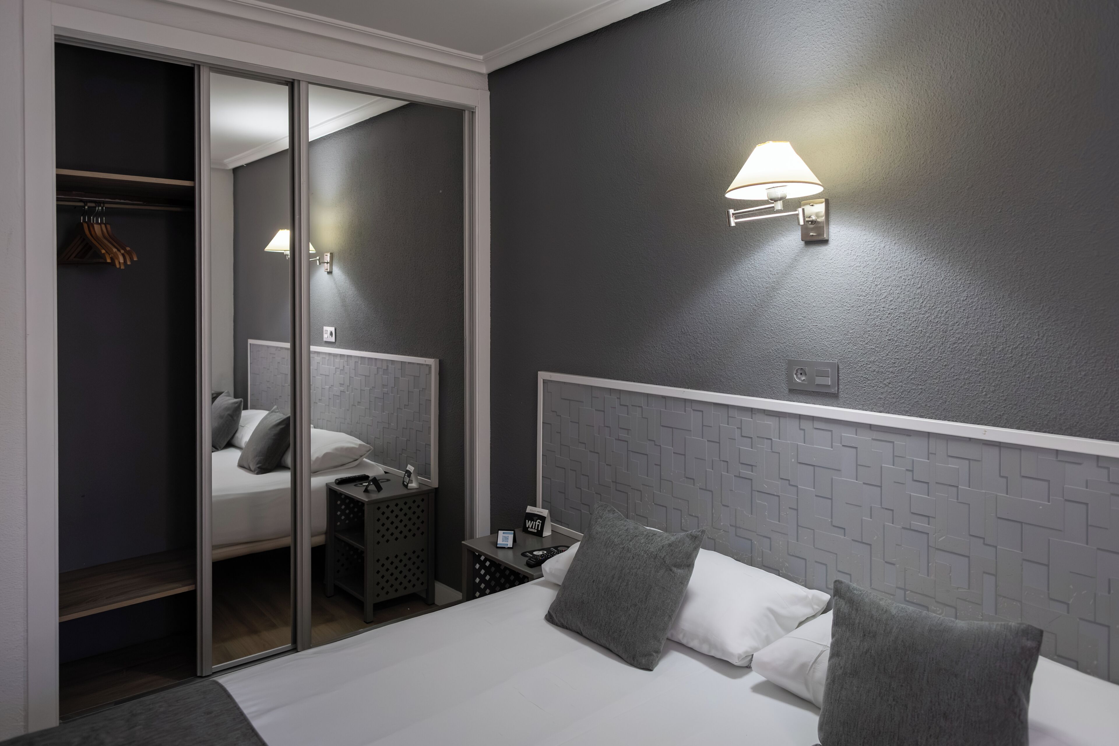 Habitación de hotel moderna con cama, mesita de noche, lámpara de pared y armario con puerta corredera.