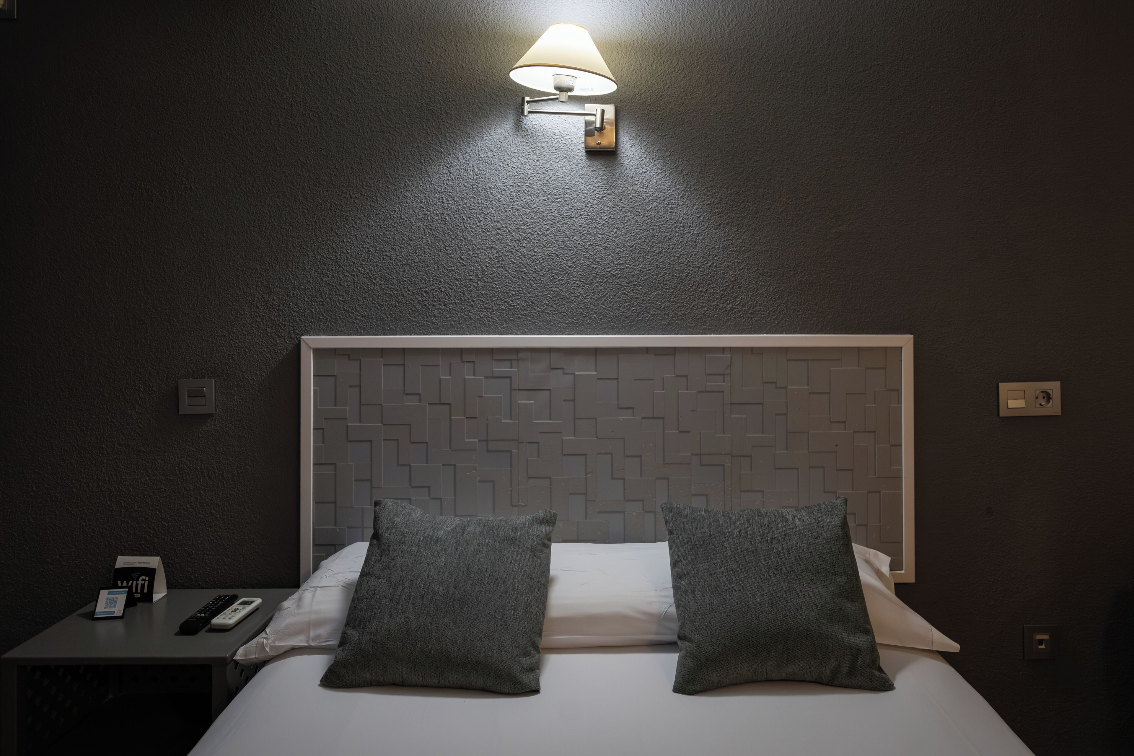 Quarto de hotel com cama, duas almofadas, mesa de cabeceira com telefone e controle remoto, e luminária fixada na parede.