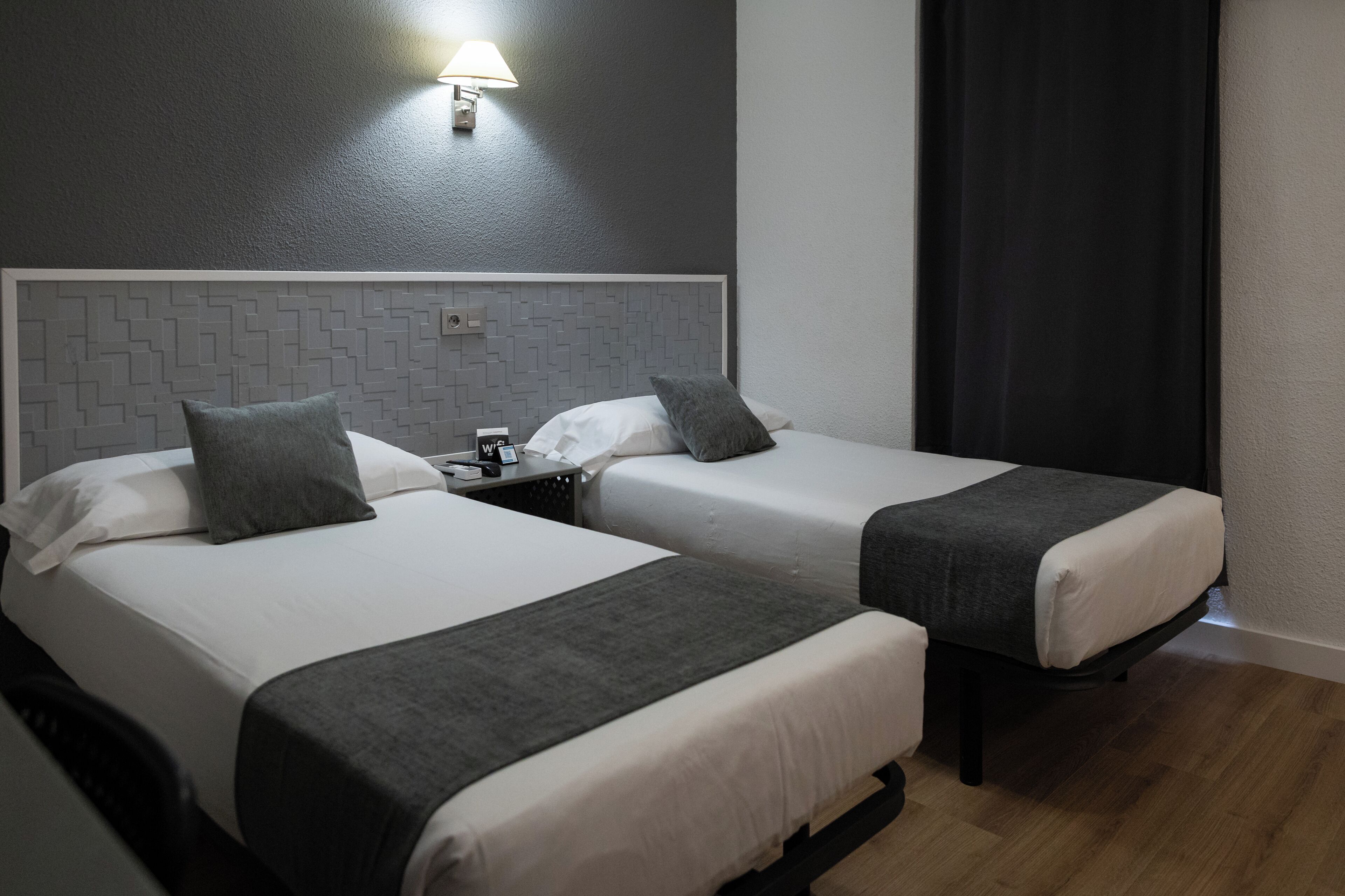 Dos camas individuales con ropa de cama gris, un cabecero con diseño geométrico, una mesita de noche con lámpara y una cortina oscura.
