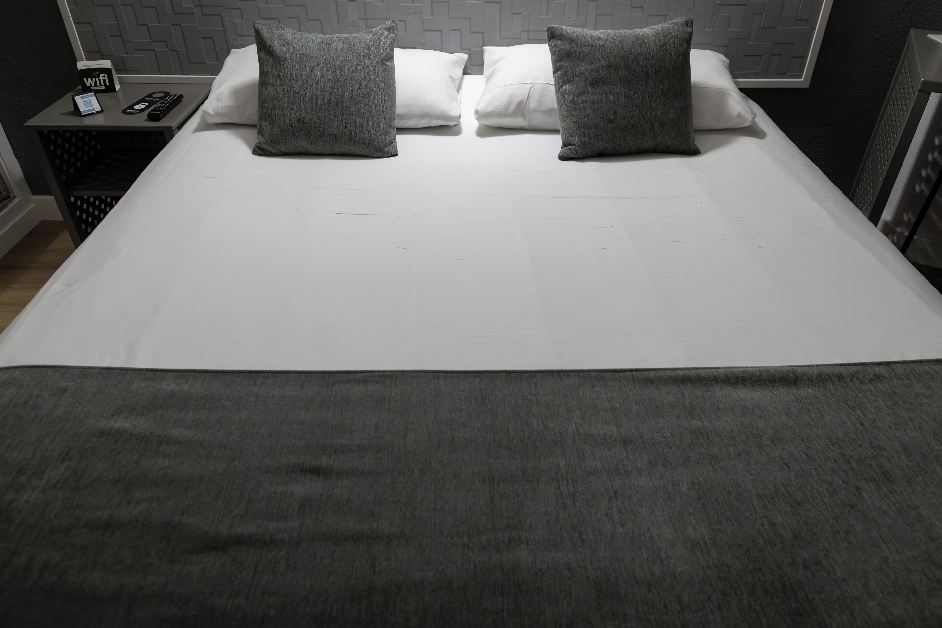 Un lit bien fait, avec du linge de lit blanc et deux oreillers gris, une petite table sur laquelle est posé un panneau indiquant la présence d’une connexion Wi-Fi, et une tête de lit ornée d’un motif géométrique.
