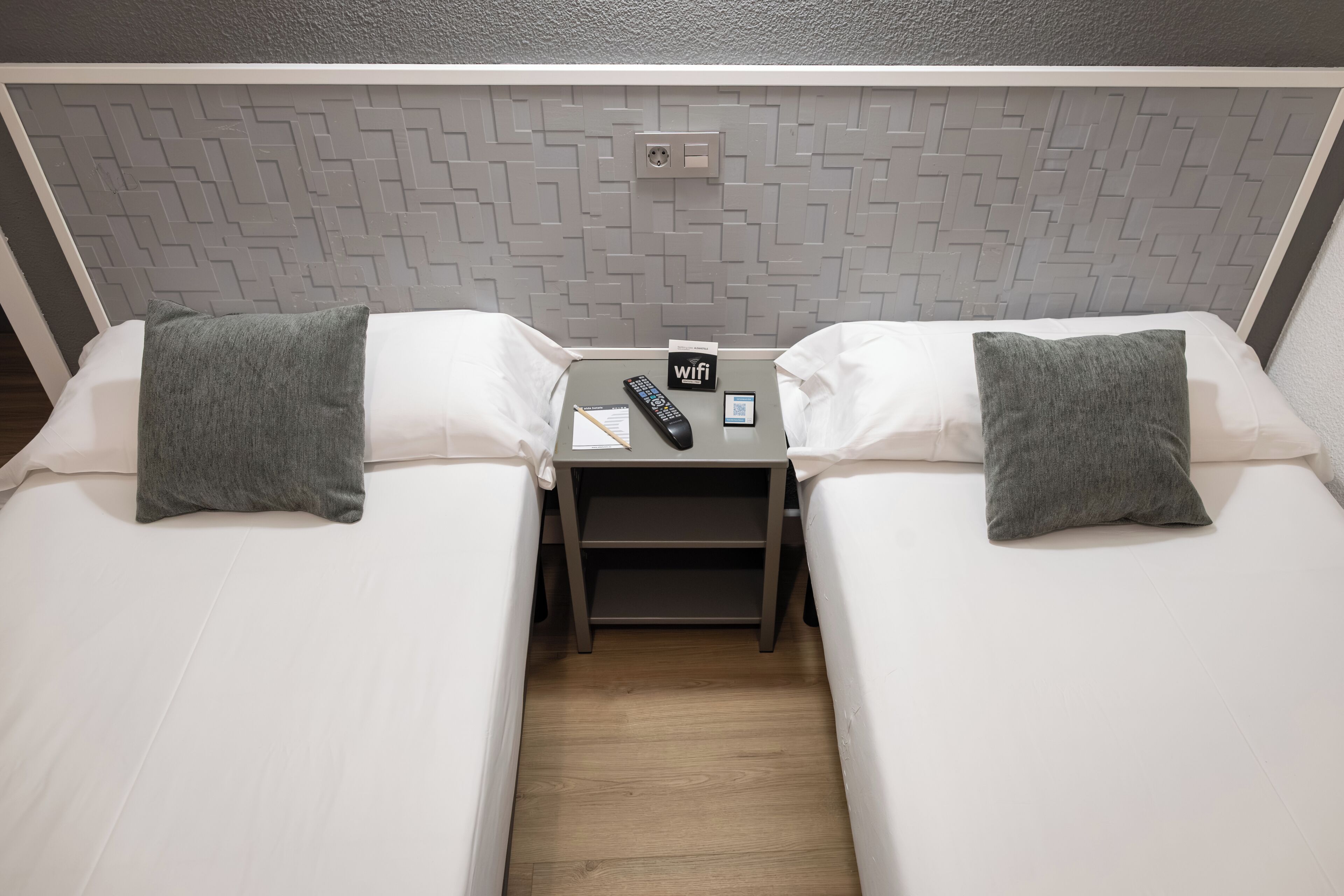A twin room with two beds, a nightstand with a remote control, a WiFi symbol, and a power outlet.