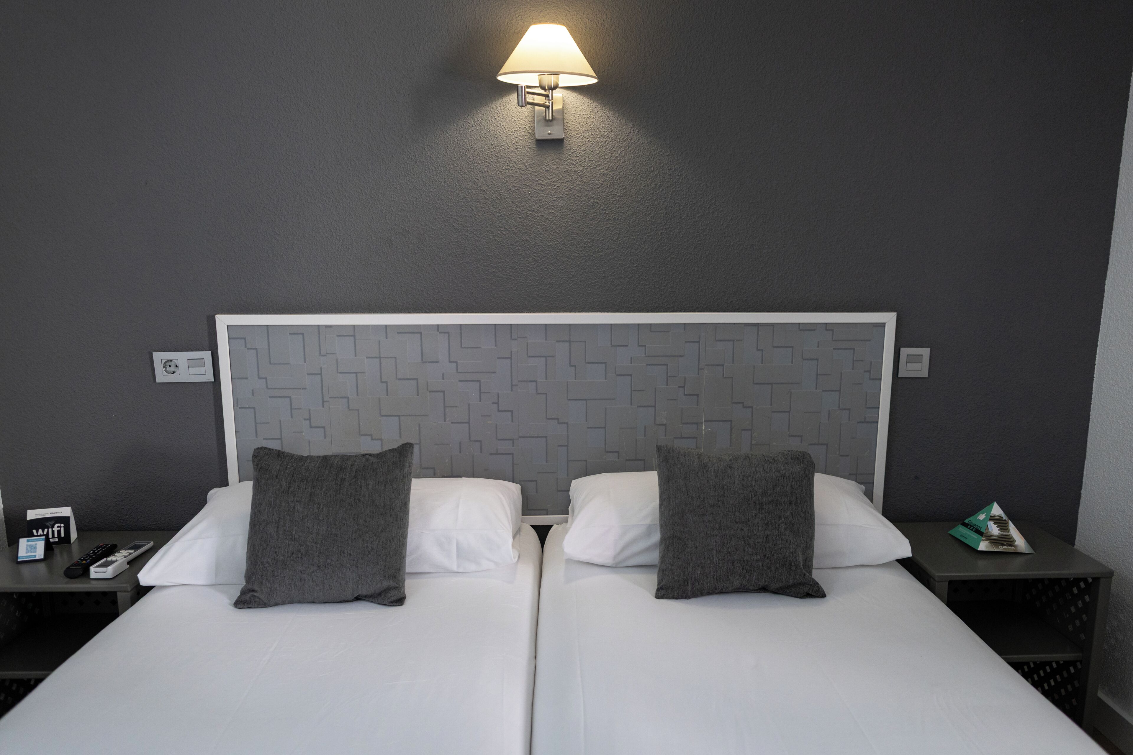 Cama doble con sábanas blancas y dos almohadas grises, lámpara fijada a la pared y mesita de noche con un control remoto y un teléfono.