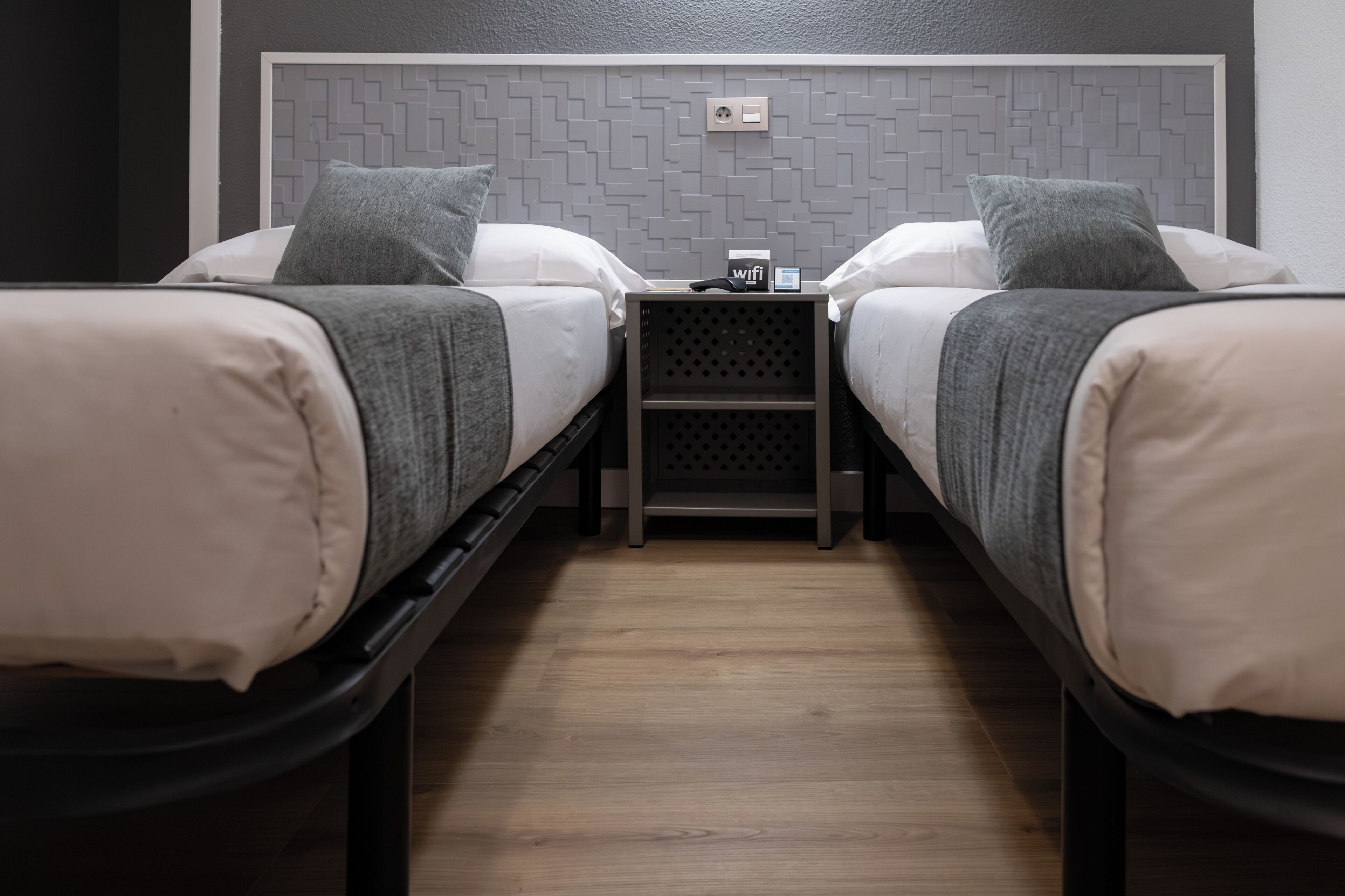 Une chambre double avec deux lits, une petite table ornée du symbole du Wi-Fi et un mur décoré d’un motif géométrique.