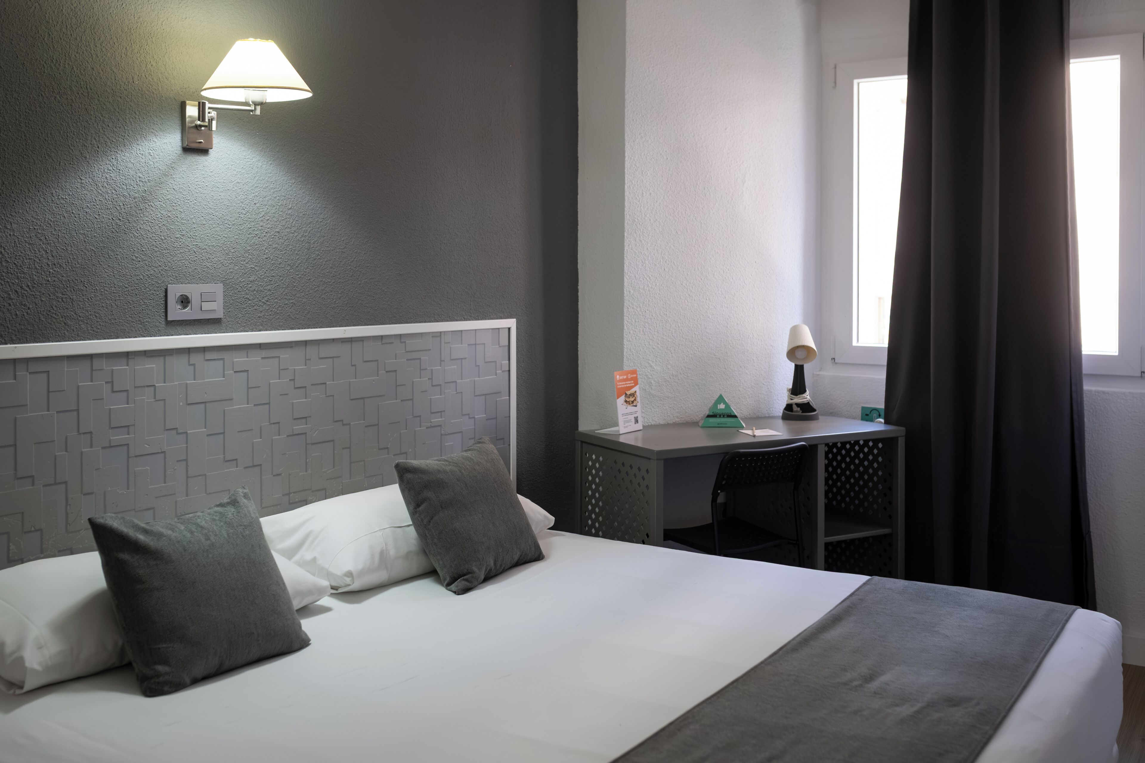 A hotel room with a bed, a desk, a lamp, and a window with curtains.