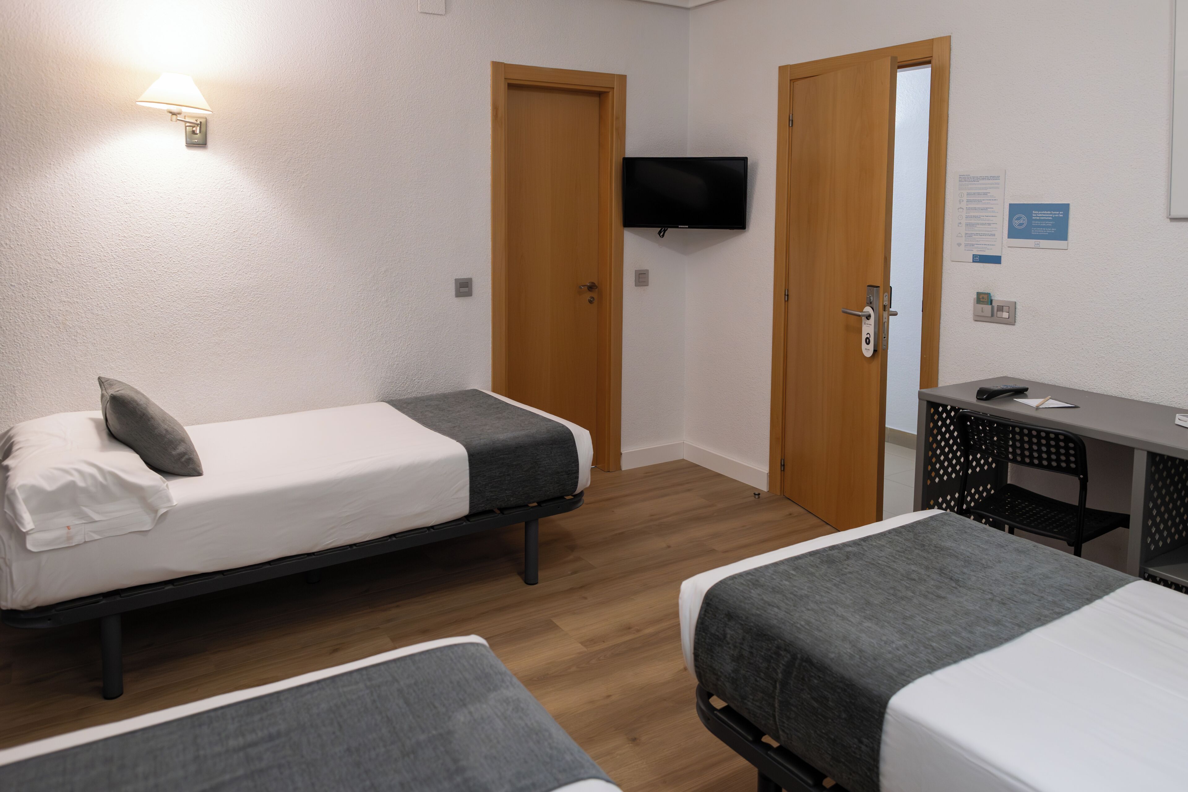A room with two single beds, a desk with a chair, a TV mounted on the wall, and a door.