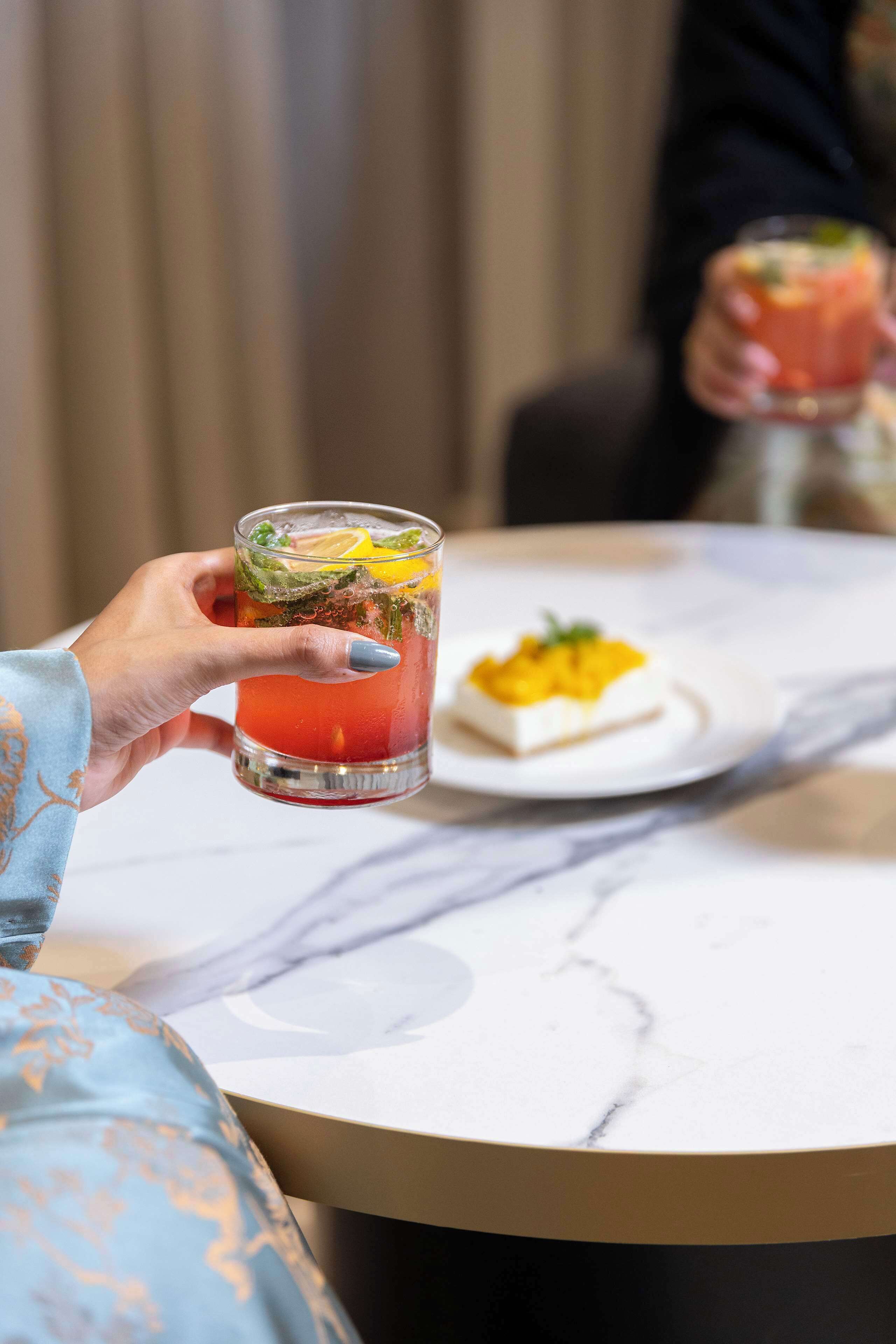 A person holding a glass of a red beverage with a garnish, a plate with a yellow dessert, and a marble table.