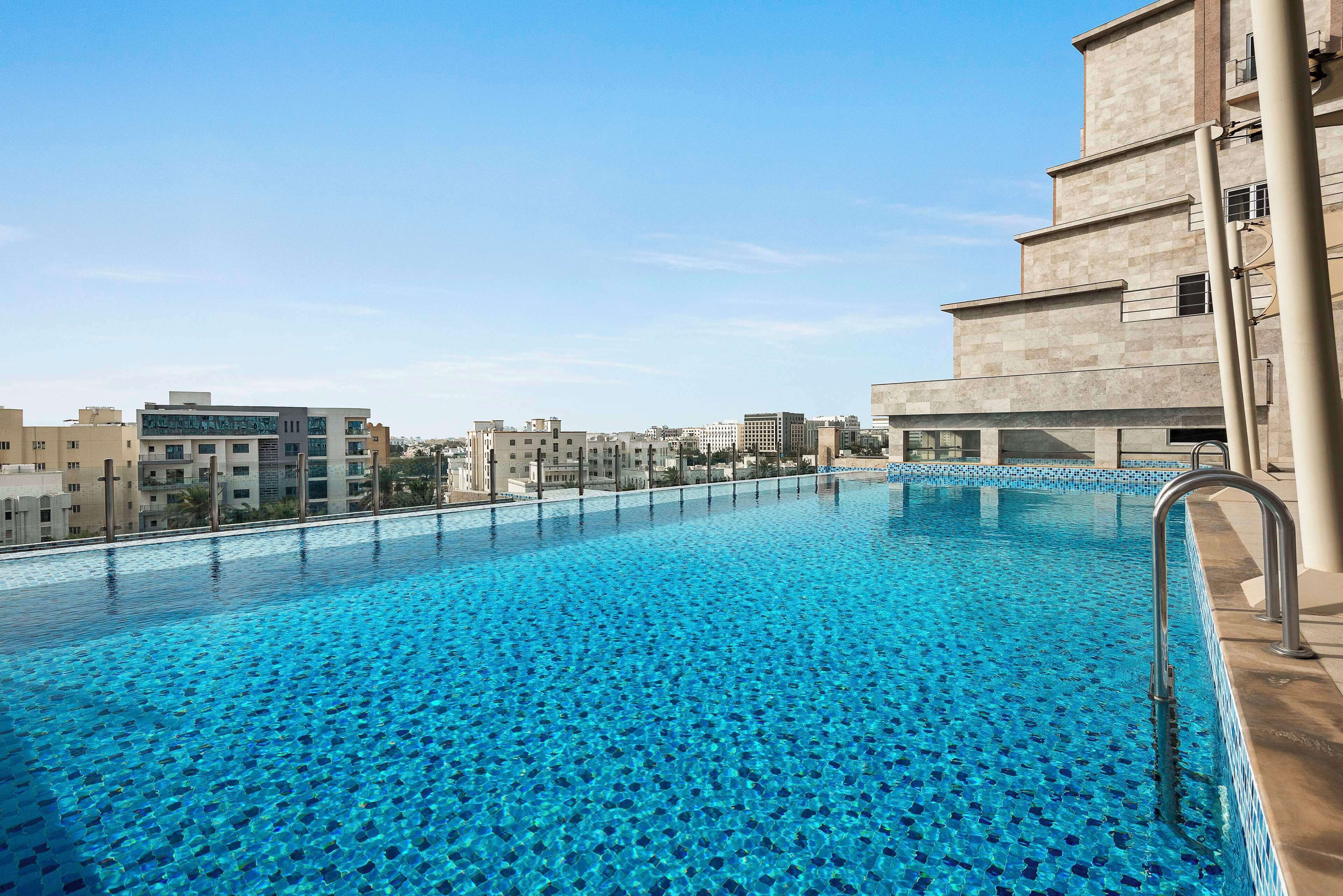A rooftop swimming pool with a clear blue tiled floor, surrounded by modern buildings.