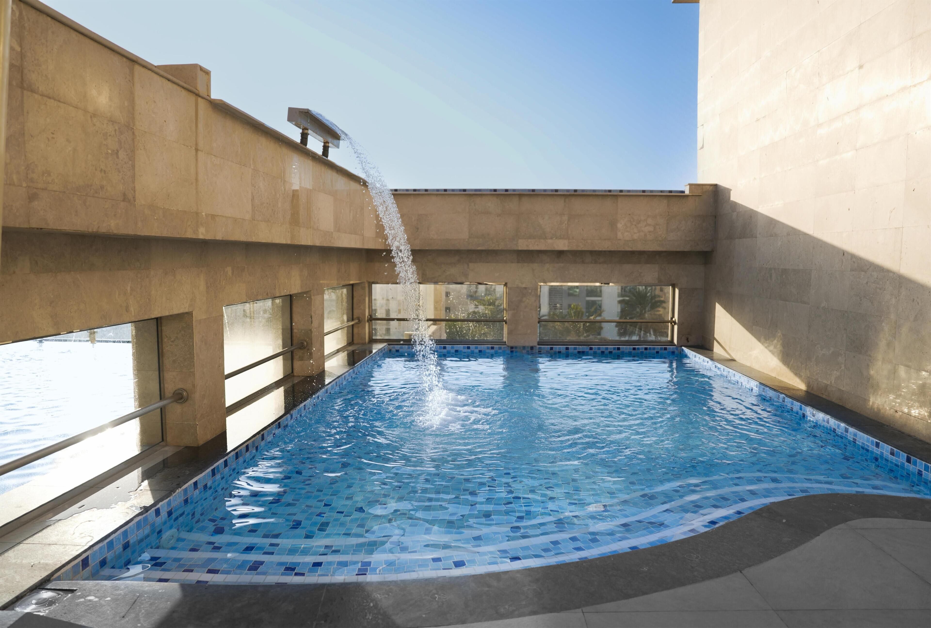 An indoor swimming pool with a clear view of the sky through large windows, a water fountain feature, and a tiled floor.