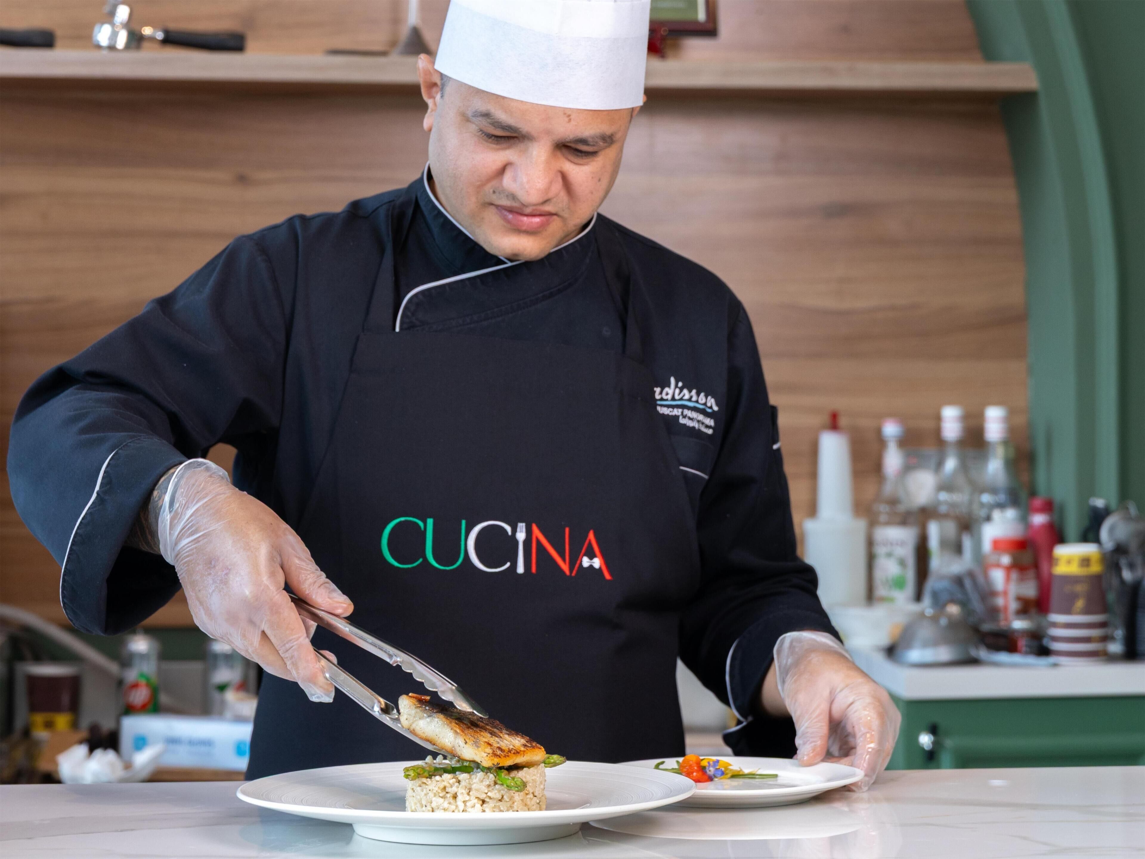 A chef in a black uniform with 'CUCINA' on the apron, preparing food on a white plate.