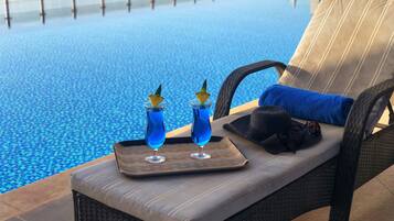 Un salon au bord de la piscine avec un fauteuil rembourré, un plateau avec deux boissons et une serviette bleue.