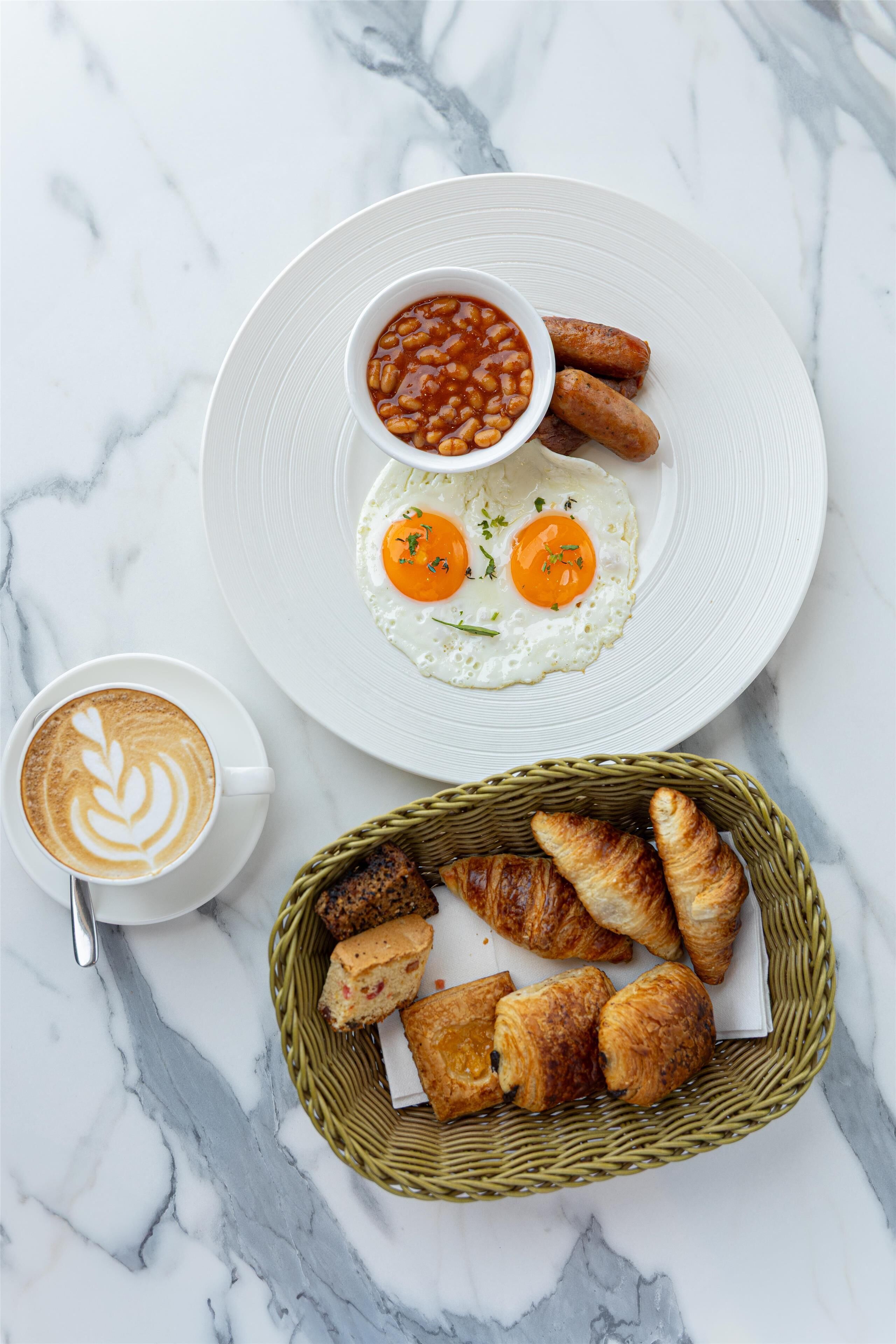 Un plato de desayuno con huevos, salchichas y frijoles, una taza de café y una cesta de pasteles sobre una superficie de mármol.
