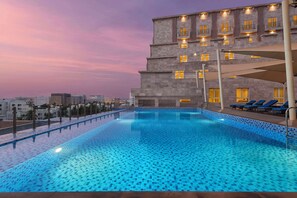 A swimming pool with a tiled edge, adjacent to a building with multiple balconies and windows, and a view of the cityscape in the background.