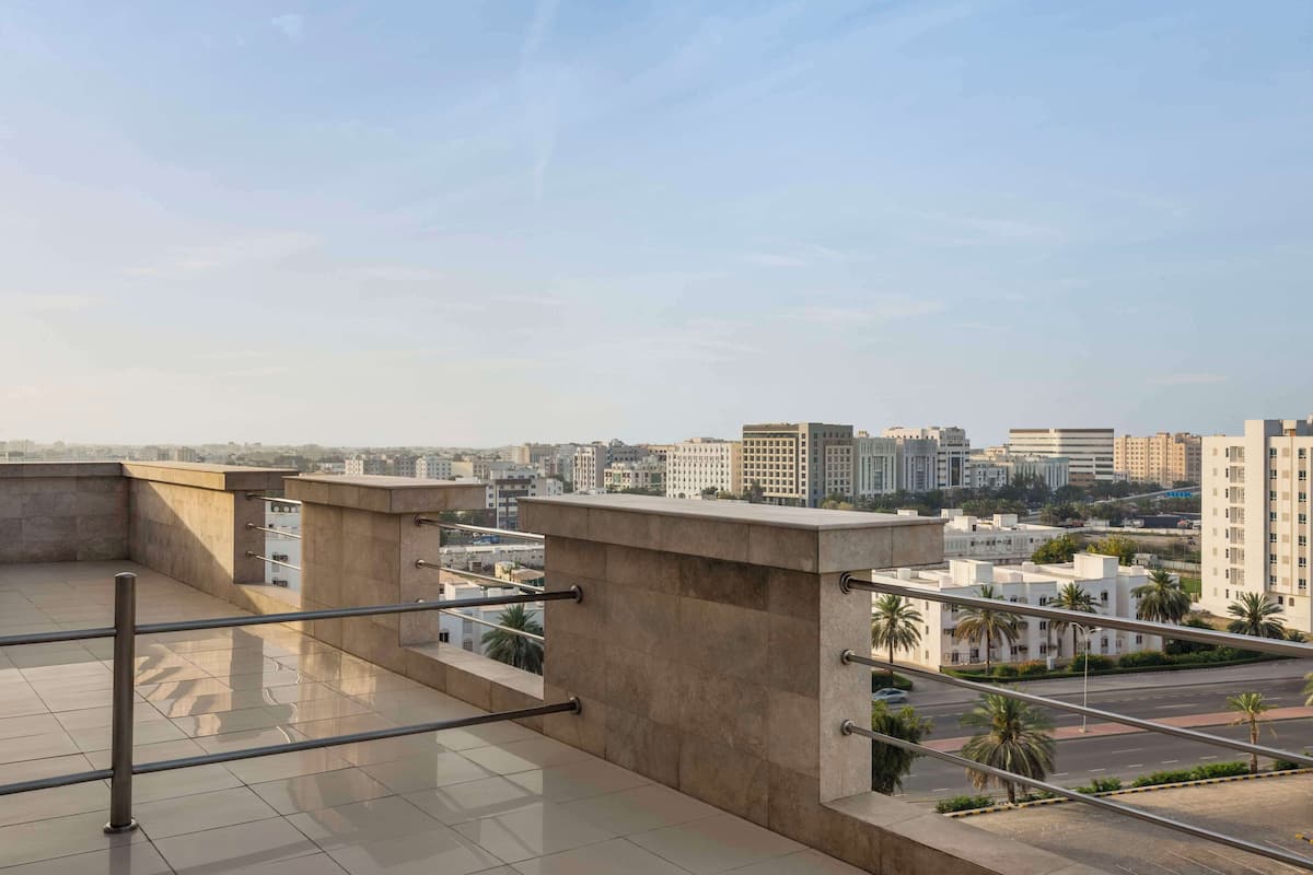 Une terrasse sur le toit, dotée d’une rambarde en métal, d’un sol en béton et offrant une vue sur un paysage urbain avec des bâtiments et des palmiers.