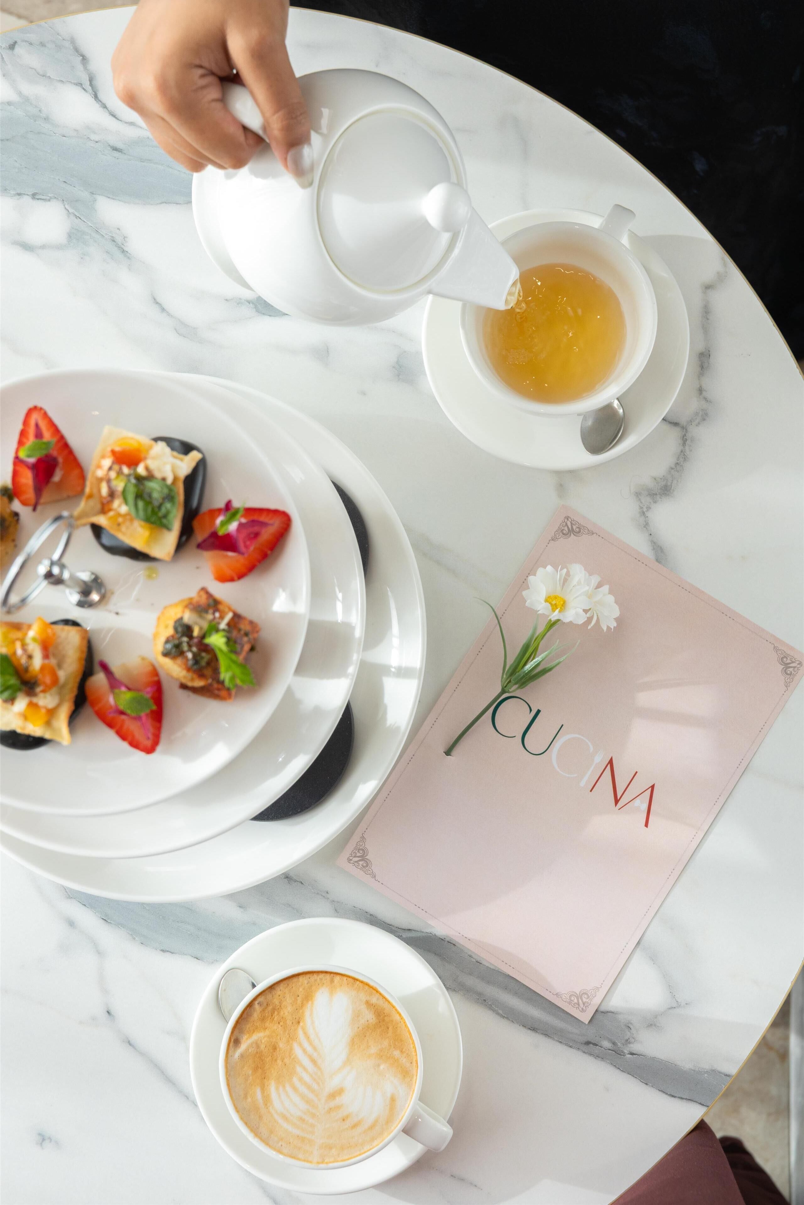 A table with a plate of food, a cup of coffee, a teapot pouring tea, and a napkin with a flower.