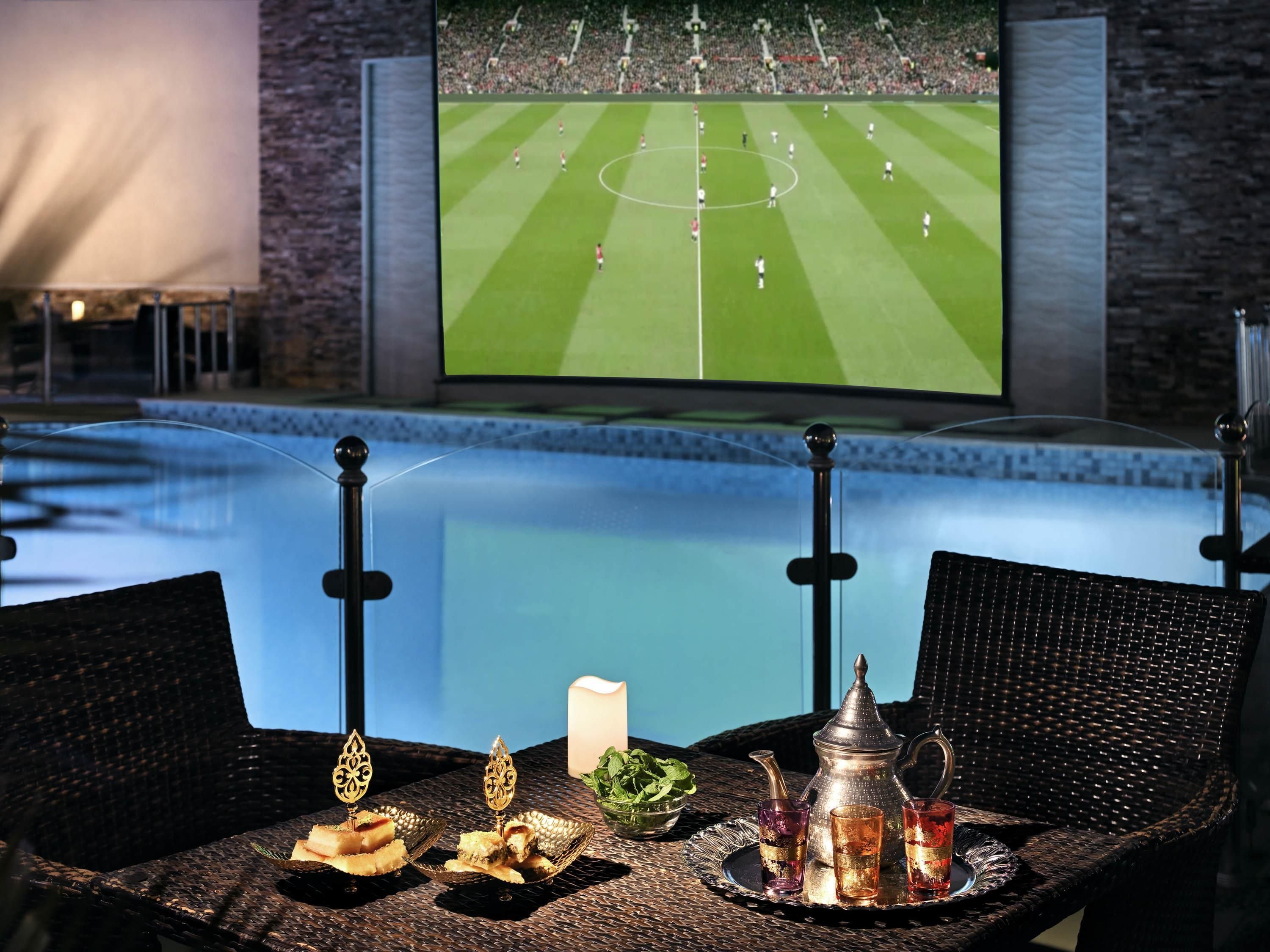 Un espace repas aménagé au bord de la piscine, avec une table dressée pour deux personnes, un téléviseur diffusant un match de football et une vue sur la piscine.