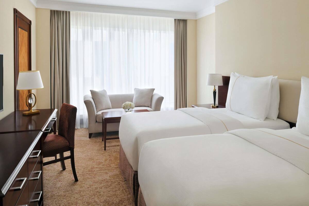 A hotel room with a bed, a desk, a chair, a small table with a vase, and a window with curtains.