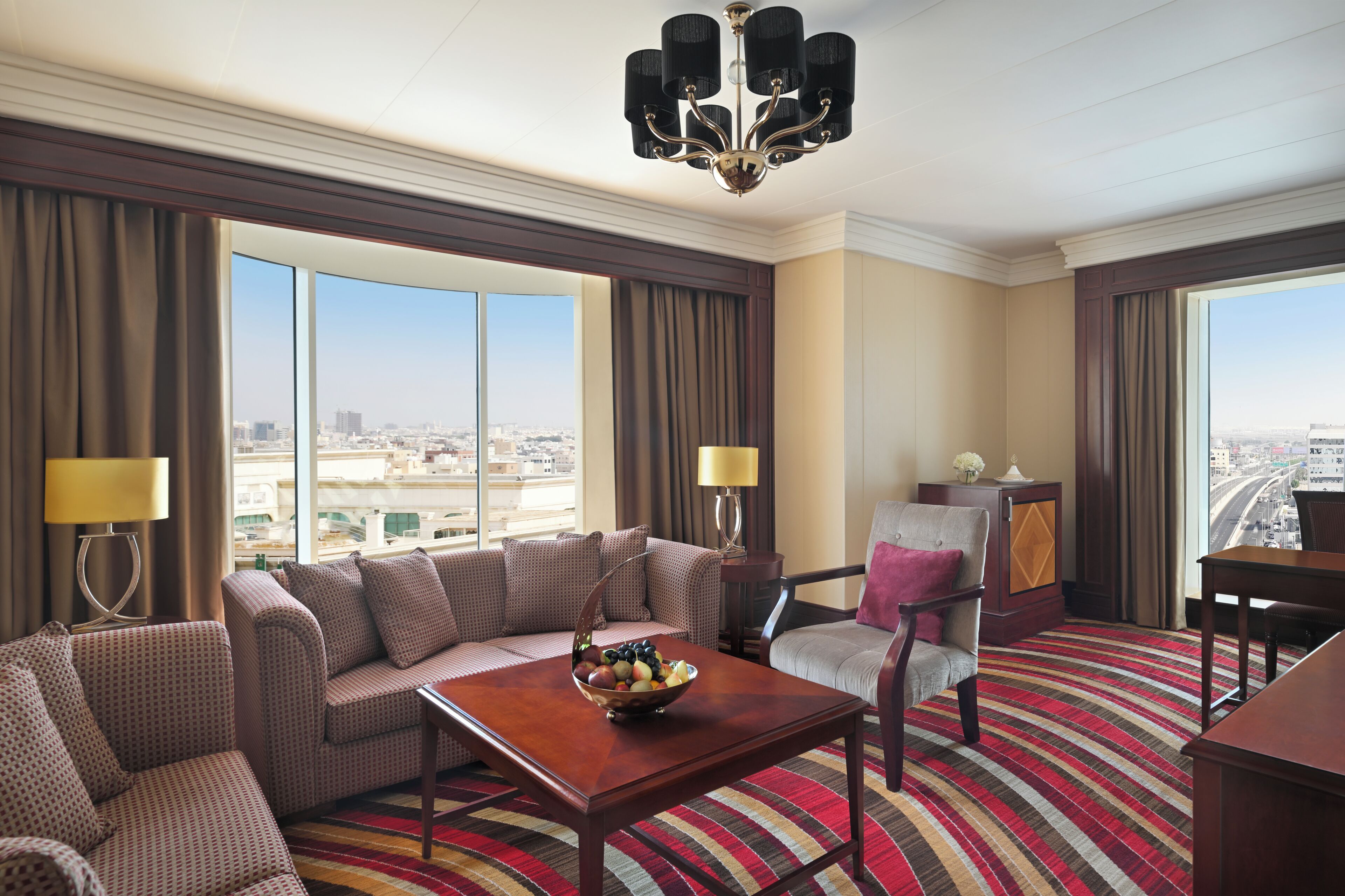 A hotel room with a large window, a sofa, a chair, a small table with a fruit bowl, and a chandelier.
