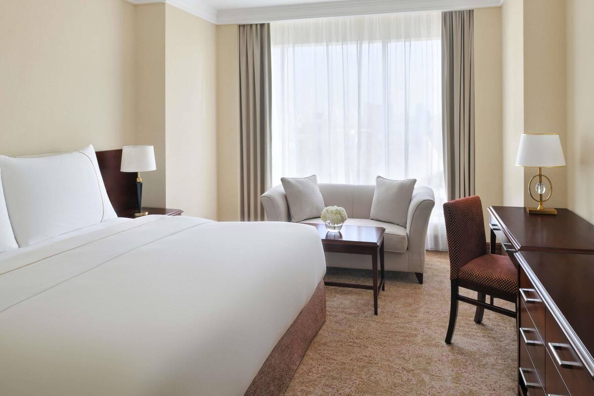 A hotel room with a large bed, a sofa, a small table with a vase, a desk with a lamp, and a chair.