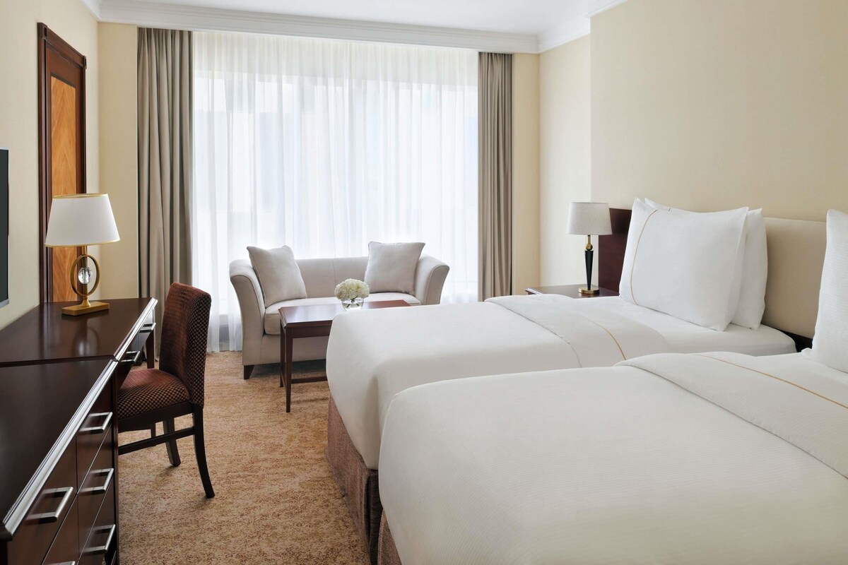 Una habitación de hotel con cama, escritorio, silla, una mesita con un jarrón y ventana con cortinas.