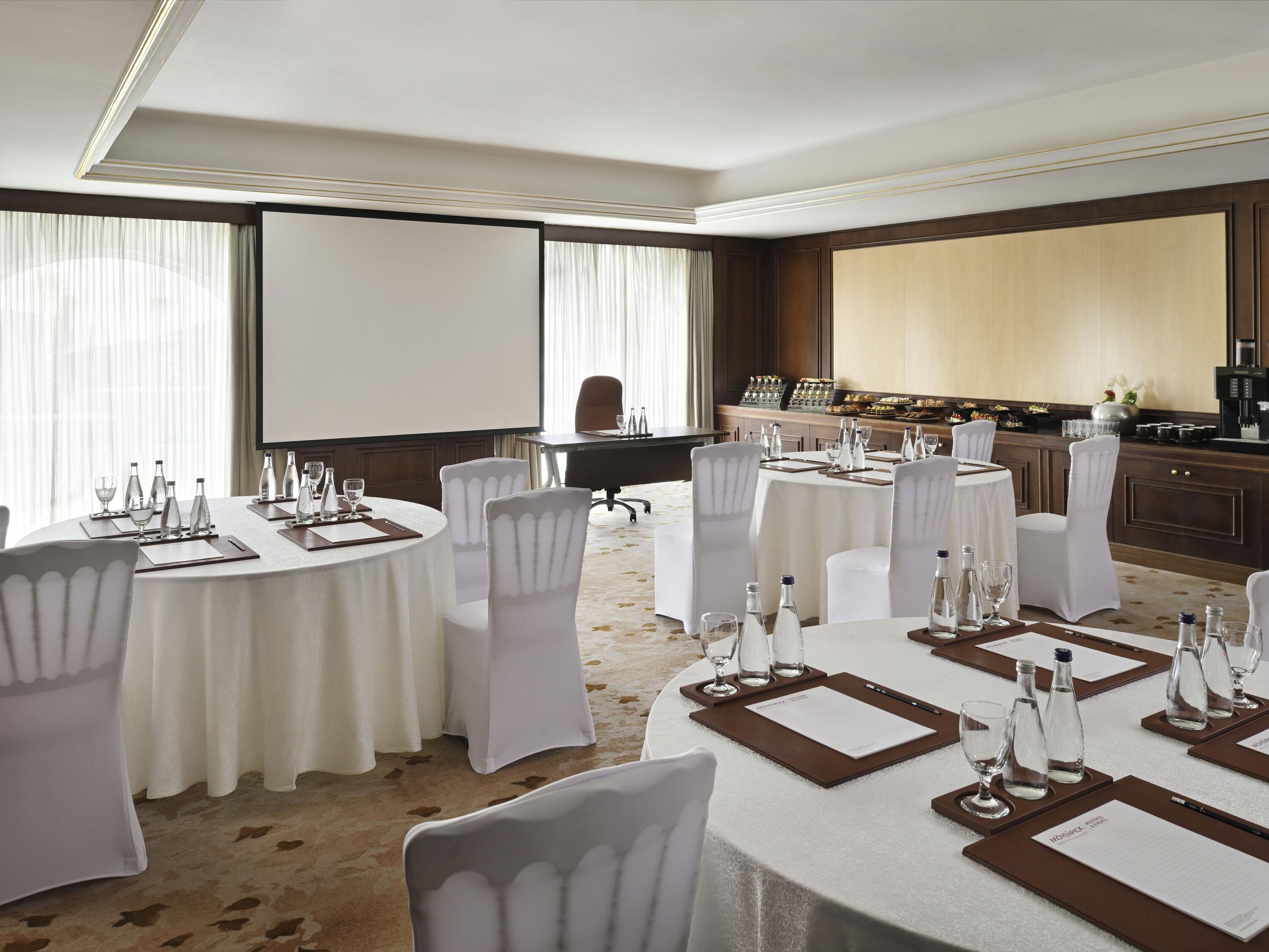 Une salle de conférence équipée de plusieurs tables rondes, de chaises blanches, d’un écran de projection et d’un coin cuisine.