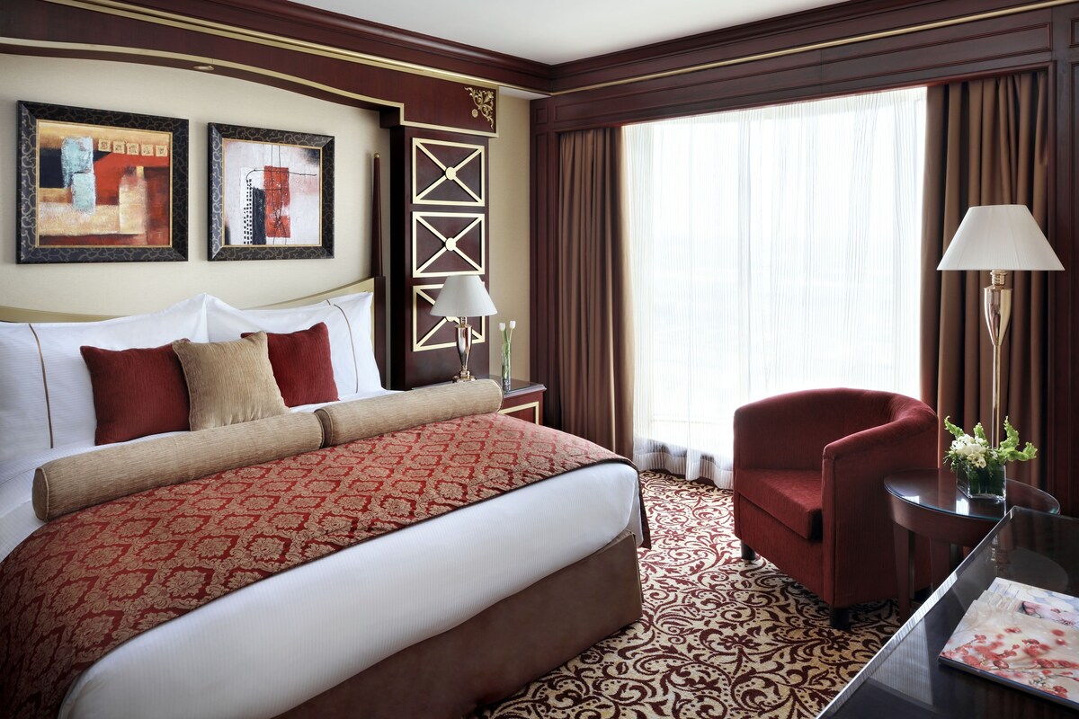 A hotel room with a large bed, a red armchair, a glass table, and two framed artworks on the wall.