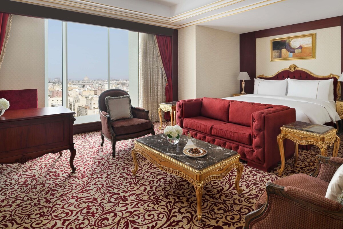 Habitación de hotel con una cama grande, un sofá rojo, una silla de madera, una mesa de centro de mármol y vistas a la ciudad.