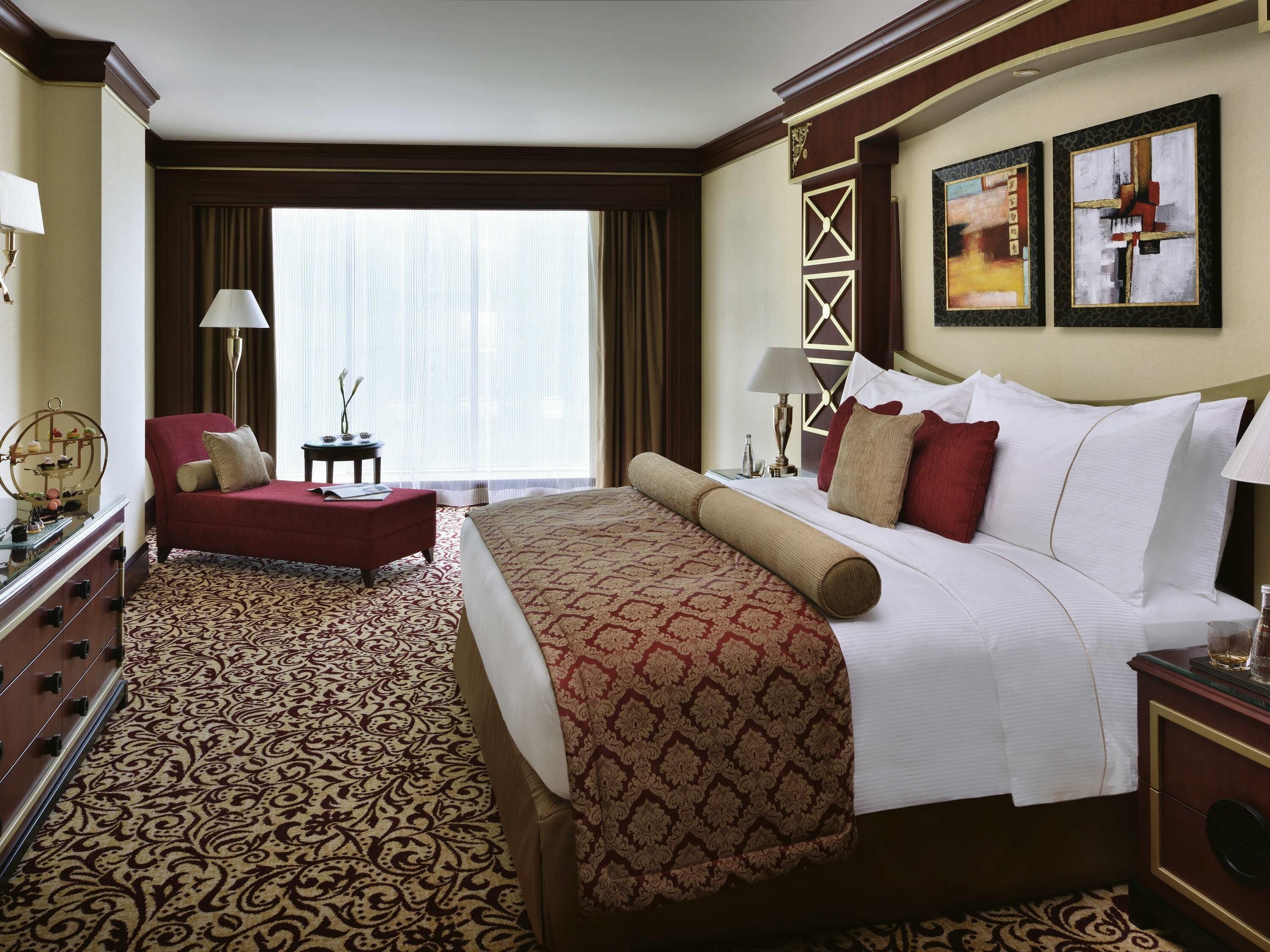 A hotel room with a large bed, a red armchair, a desk with a lamp, and a large window with curtains.