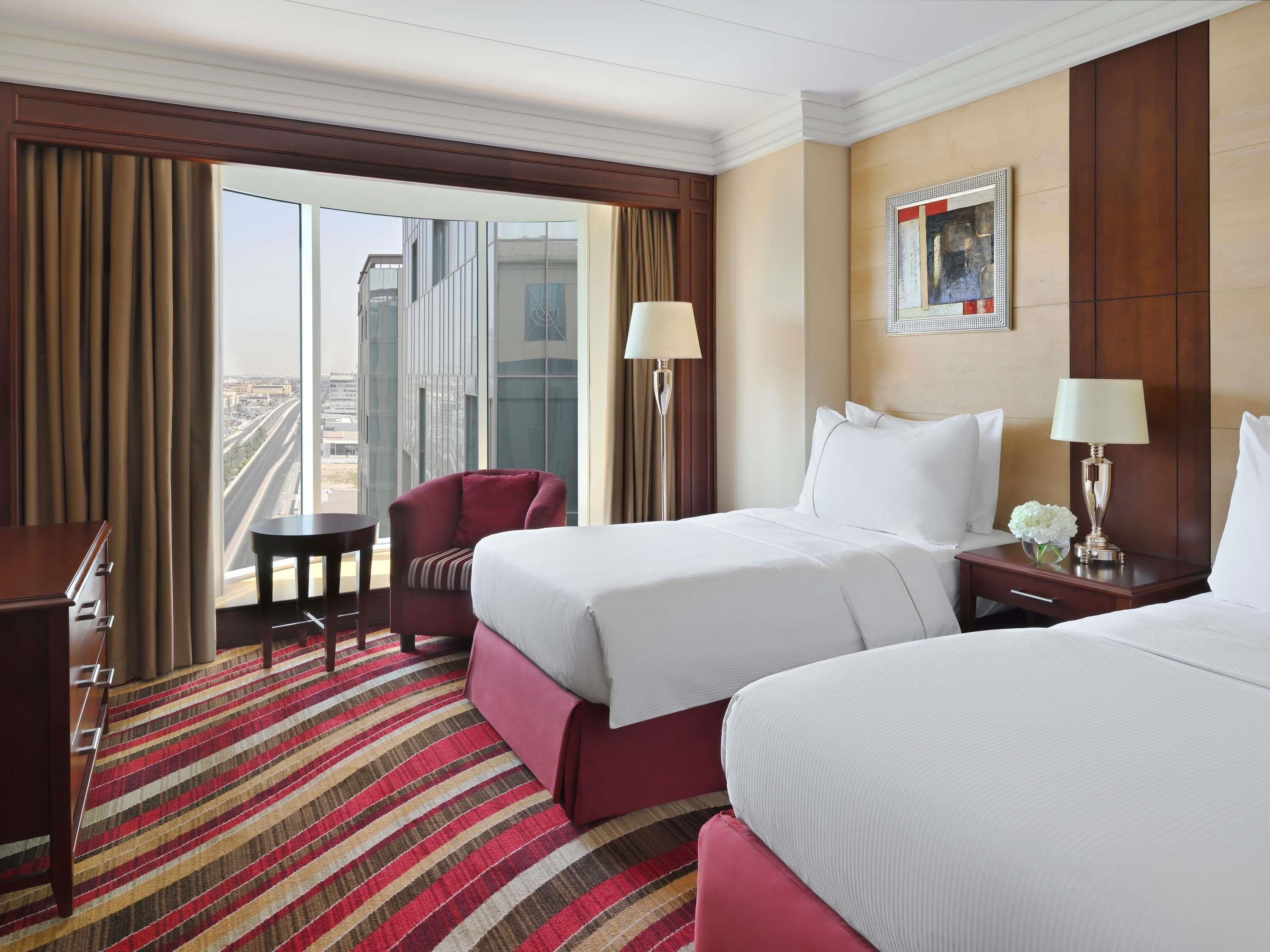 A hotel room with two beds, a desk, a chair, a window with a view of buildings, and a painting on the wall.