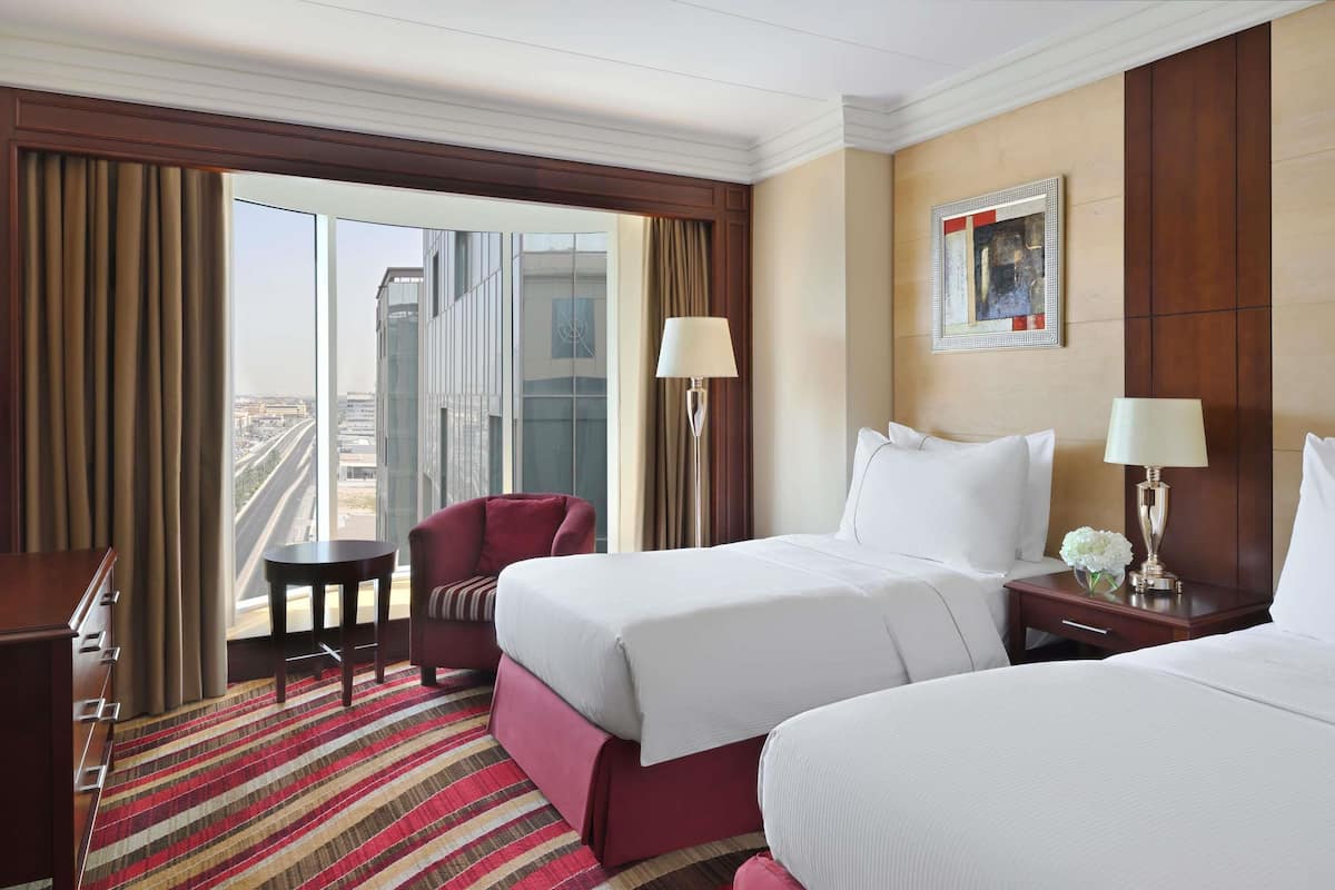 Habitación de hotel con dos camas, un escritorio, una silla, una ventana con vista a edificios y un cuadro en la pared.