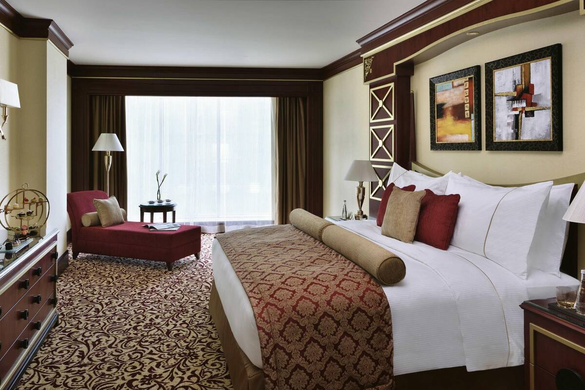 Una habitación de hotel con una cama grande, un sillón rojo, un escritorio con lámpara y un ventanal con cortinas.