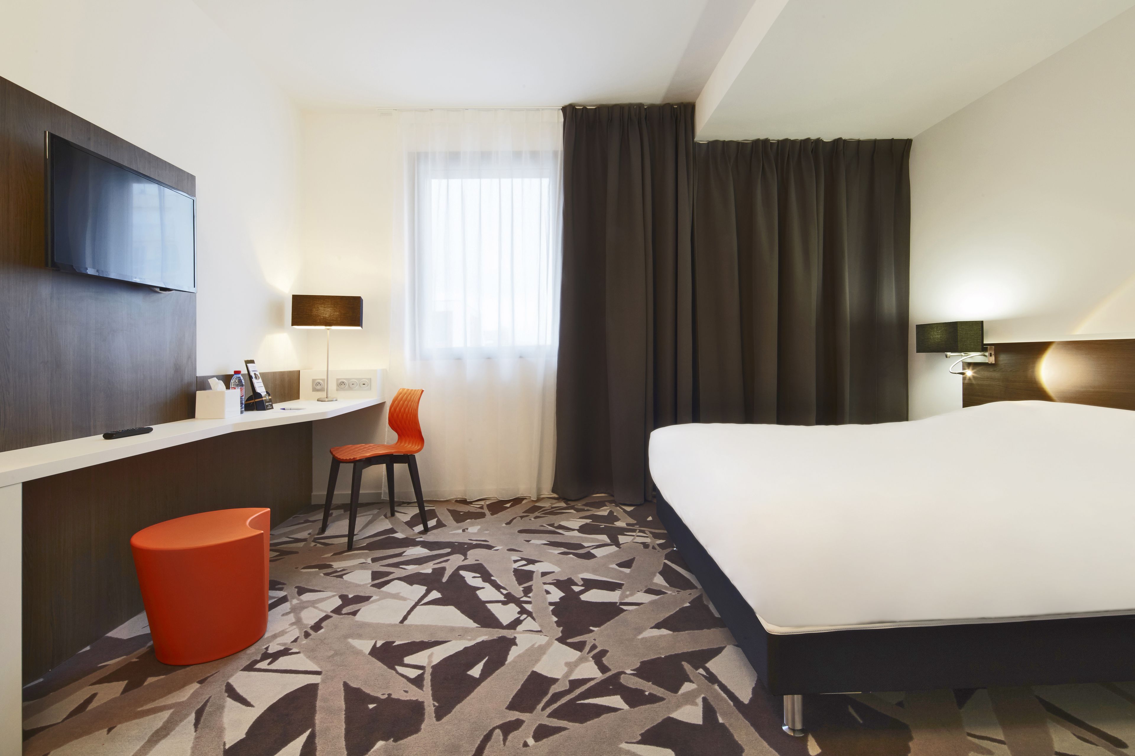 A hotel room with a bed, a desk, a chair, a TV, and a window with curtains.