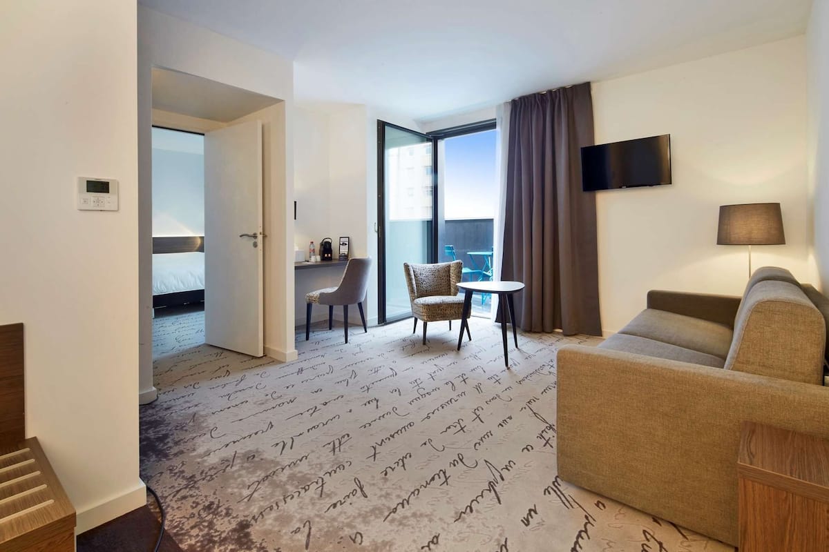 A hotel room with a sofa, a chair, a small table, a TV, and a balcony with a view.