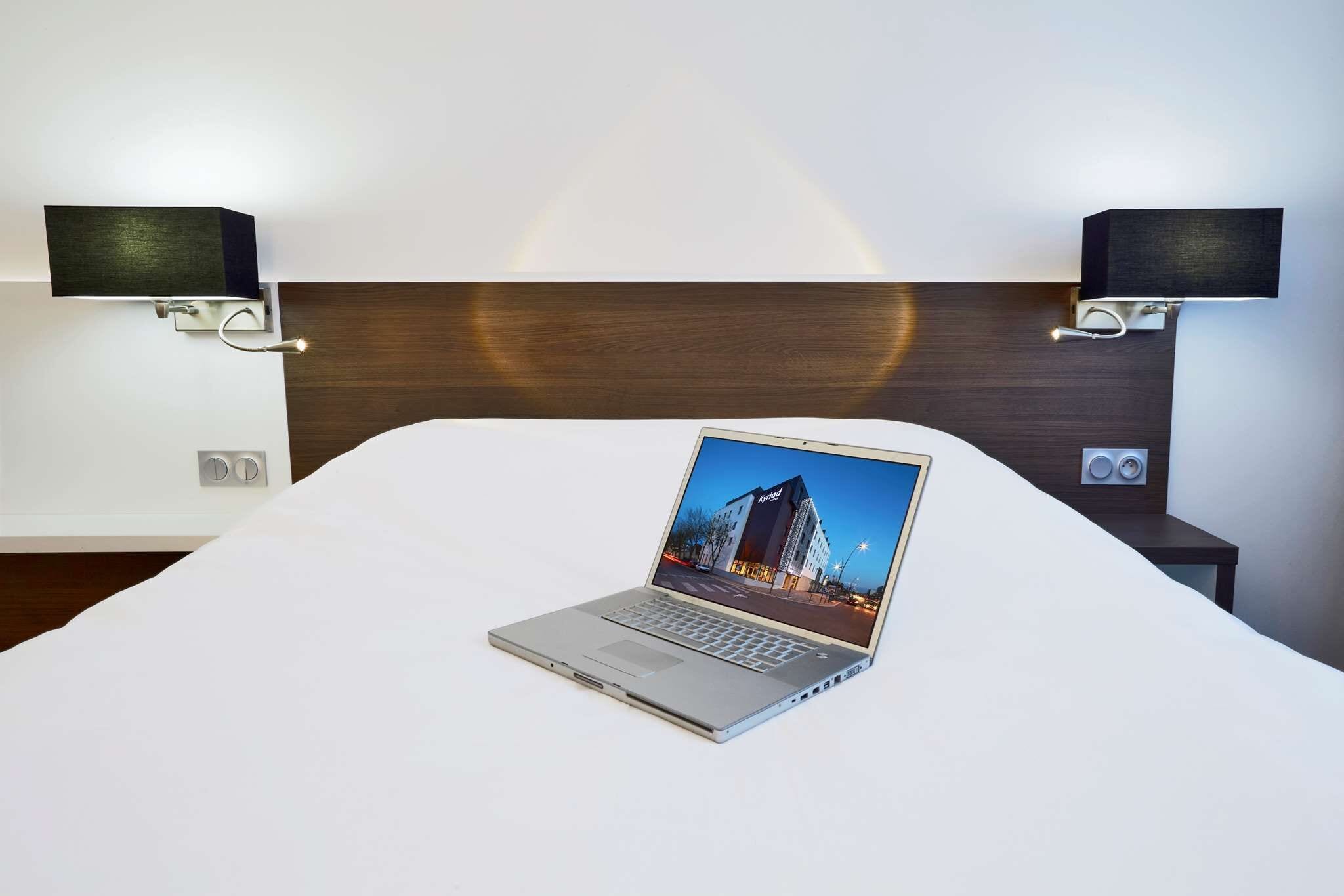 Quarto de hotel com cama, um laptop sobre a cama e dois alto-falantes fixados na parede.