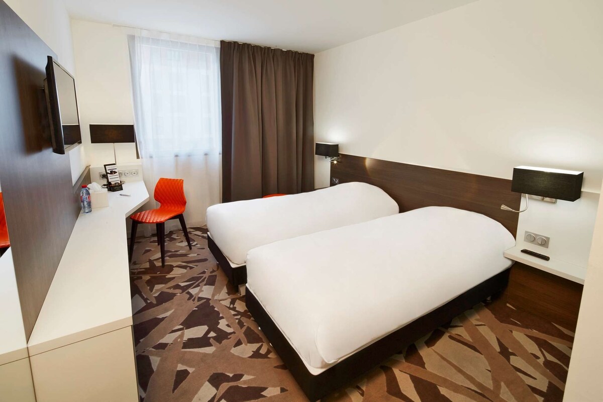 A hotel room with two beds, a desk with a chair, a TV, and a window with curtains.