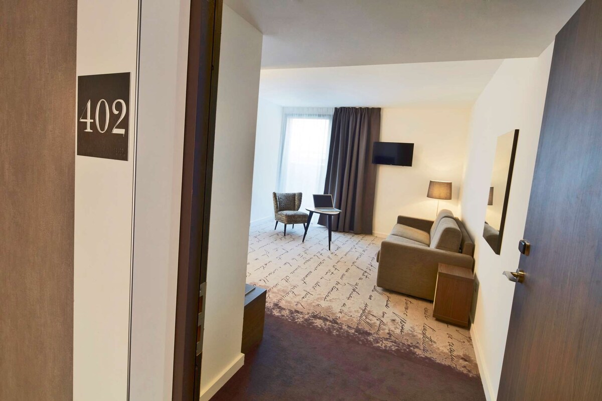A hotel room with a number 402 sign, a sofa, a chair, a small table, and a TV.