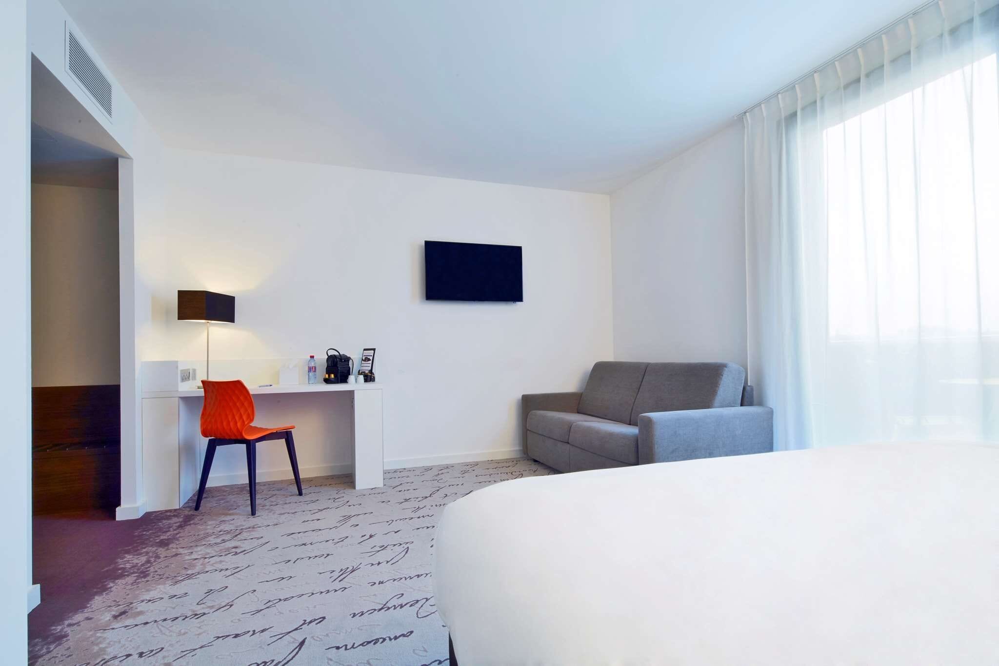 A hotel room with a bed, a desk with a chair, a sofa, a wall-mounted TV, and a window with sheer curtains.