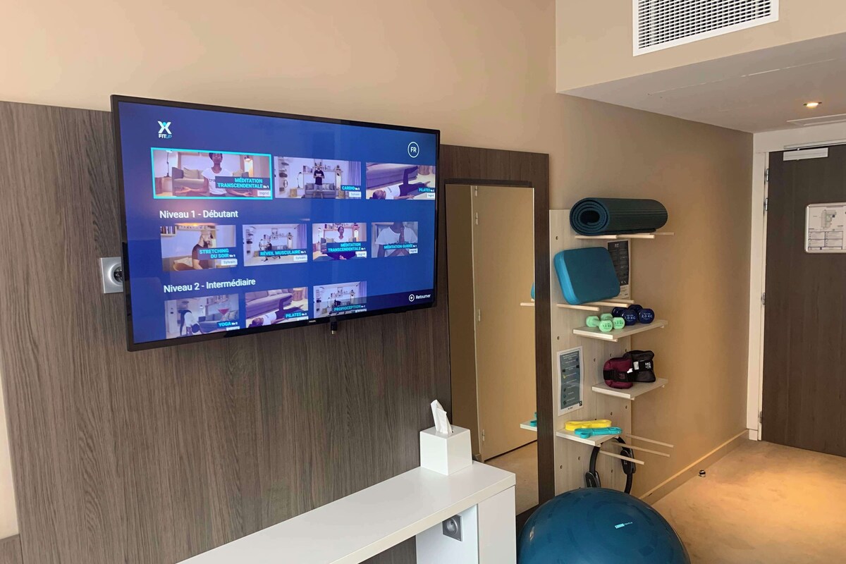 A fitness center with a wall-mounted TV displaying various exercise classes, a blue exercise ball, and a shelf with fitness equipment.