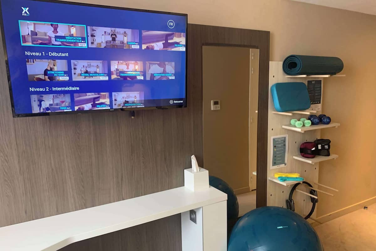 A fitness room with a wall-mounted TV displaying exercise videos, a blue exercise ball, and various fitness equipment on shelves.