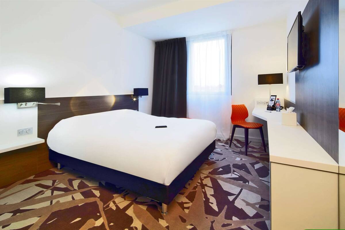 A hotel room with a large bed, a desk, a chair, a TV, and a window with curtains.