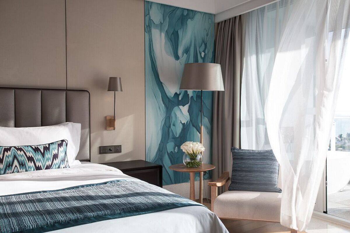 Une chambre d’hôtel moderne avec un grand lit, une table de chevet avec une lampe, une chaise et une vue sur la ville à travers de fines rideaux.
