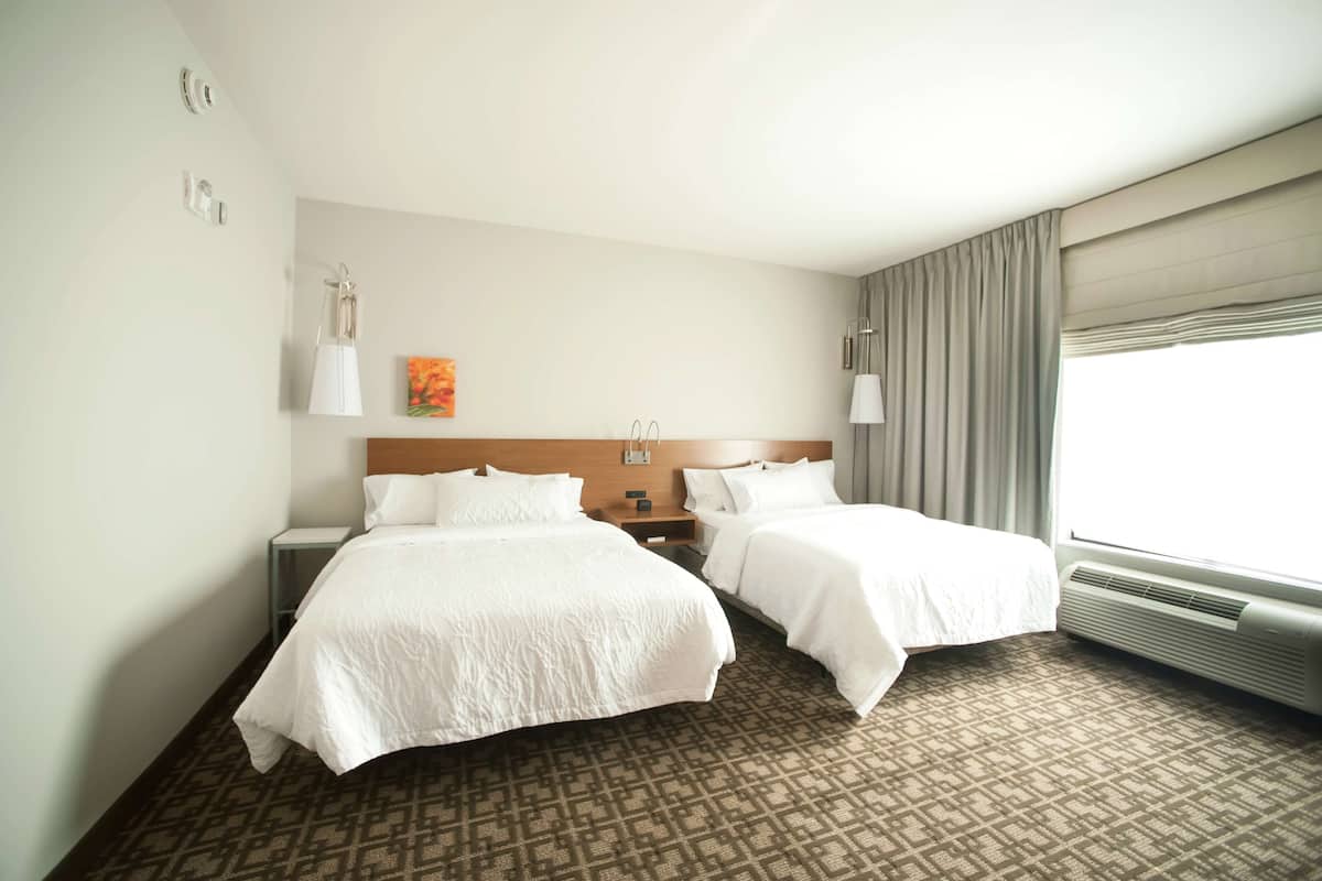 Habitación de hotel con dos camas, una mesita de noche, una lámpara, un cuadro y una ventana con cortinas.