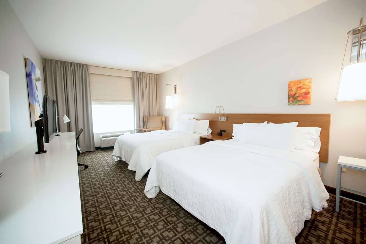 Habitación de hotel con dos camas, un escritorio, una silla, un televisor y un ventanal con cortinas.