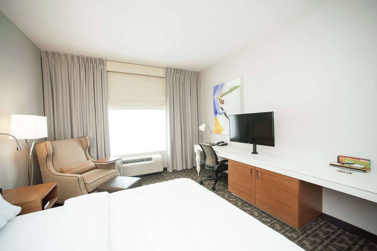 Una habitación de hotel con una cama, un escritorio con computadora, una silla, una lámpara y una ventana con cortinas.