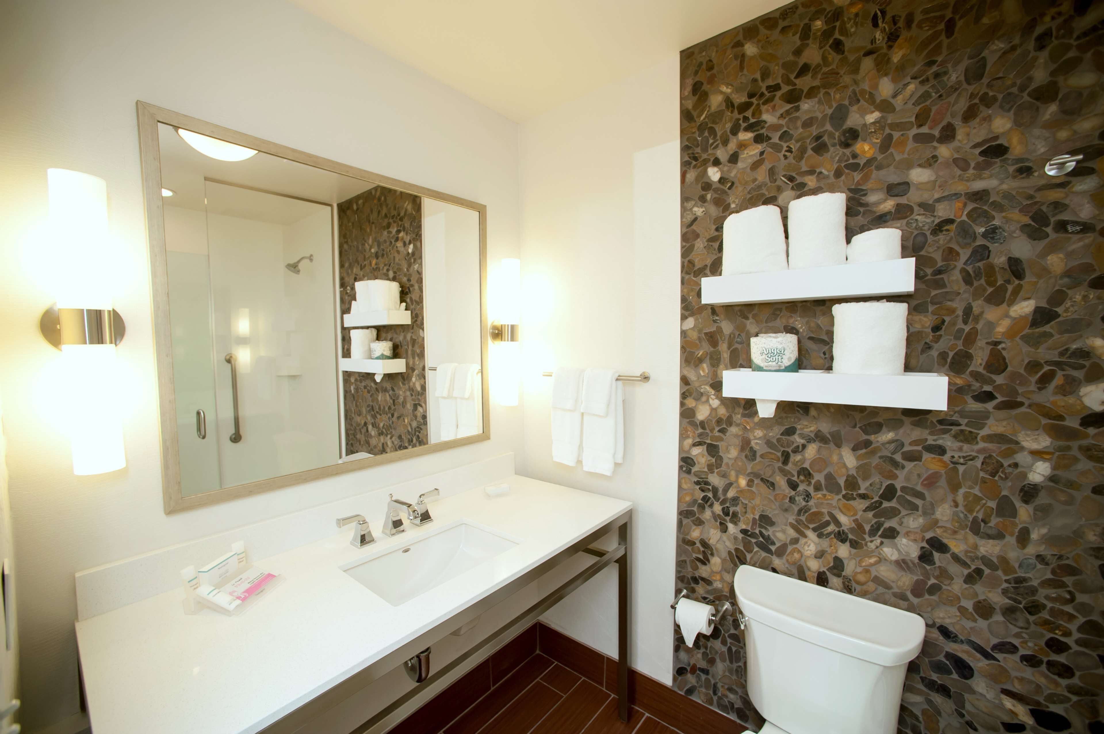 A bathroom with a white sink, a large mirror, and a stone wall. There are shelves with towels and toiletries.