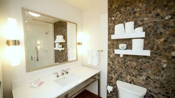 A bathroom with a white sink, a large mirror, and a stone wall. There are shelves with towels and toiletries.