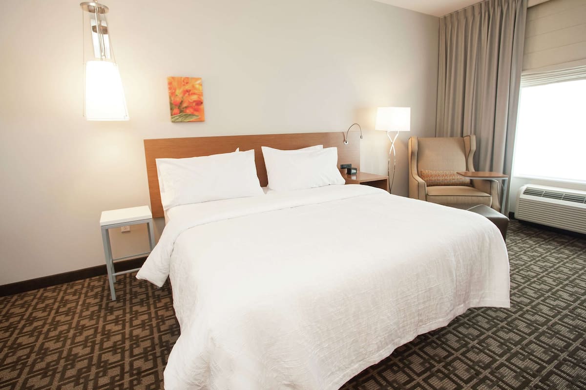 Habitación de hotel con una cama grande, una silla, una mesita de noche, una lámpara y un cuadro en la pared.
