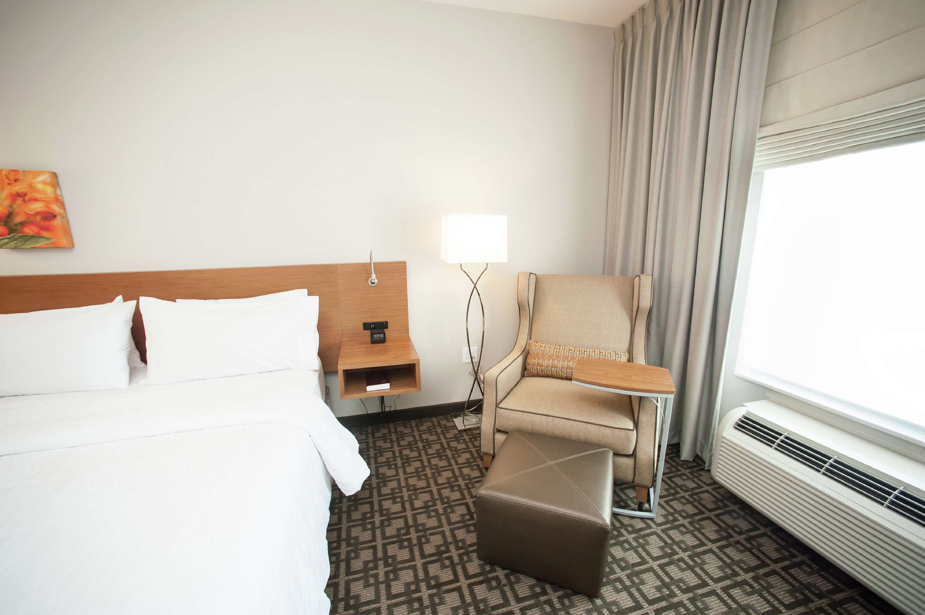 A hotel room with a bed, a chair, a small table, a lamp, and a window with curtains.