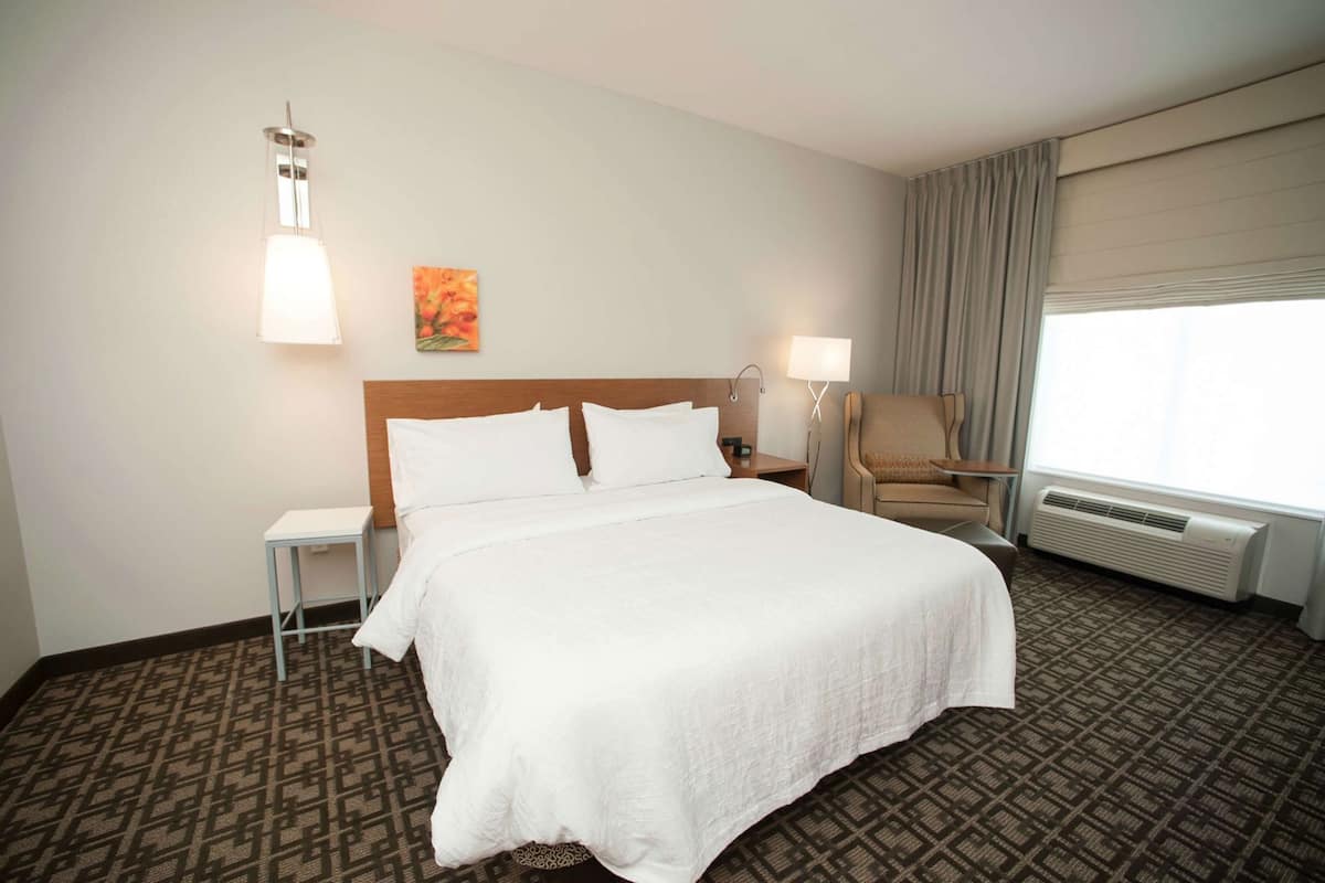 Habitación de hotel con cama, mesitas de noche, una silla, lámpara, un cuadro y ventana con cortinas.