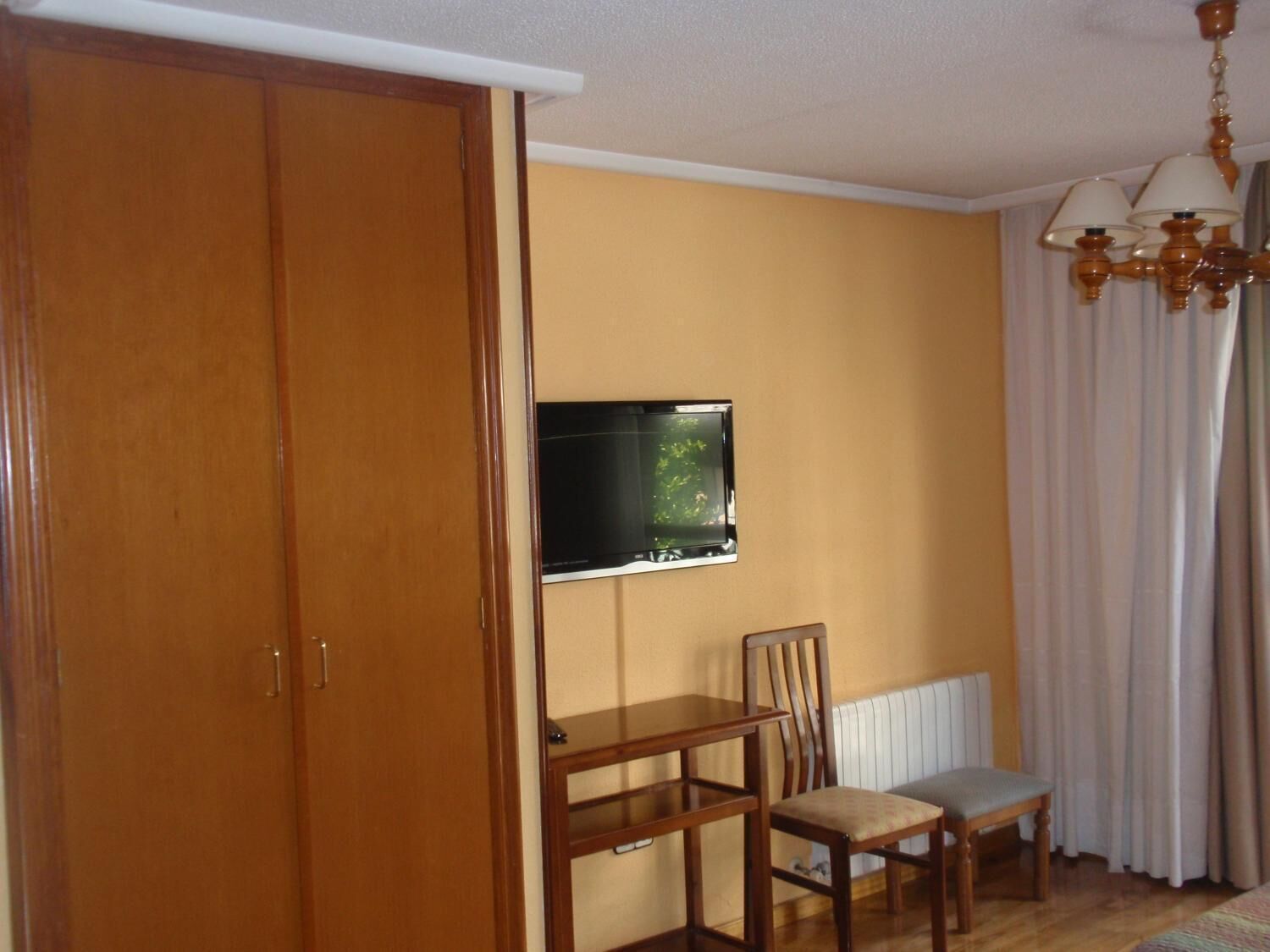 A room with a wooden wardrobe, a television mounted on the wall, a wooden table with a chair, and a radiator.