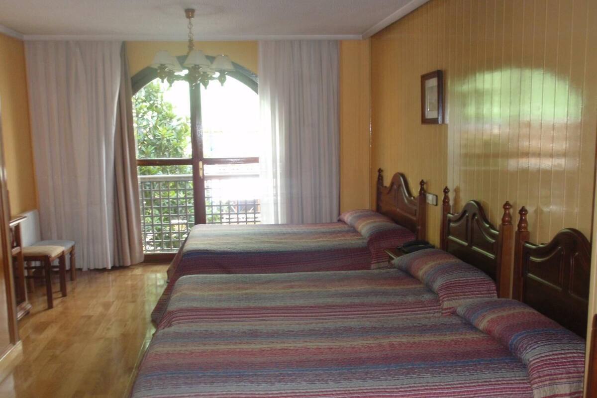 Une chambre d’hôtel avec deux lits, une tête de lit en bois, un petit bureau avec une chaise et une fenêtre avec des rideaux.